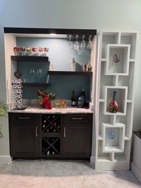 Compact home bar with dark cabinets, marble counter, wine storage, and white wall shelves with decor