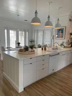Bright white kitchen with large island, pendant lights, wooden floor, and potted plants on the counters.