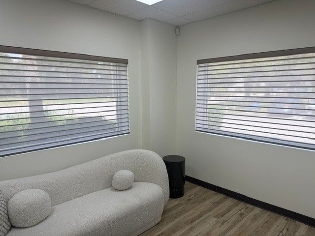 Small office with a white sofa, blinds on two windows, and a black trash can in the corner.
