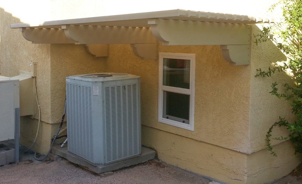 A light-colored outdoor HVAC unit sits beneath a beige, wall-mounted wooden overhang next to a small window.