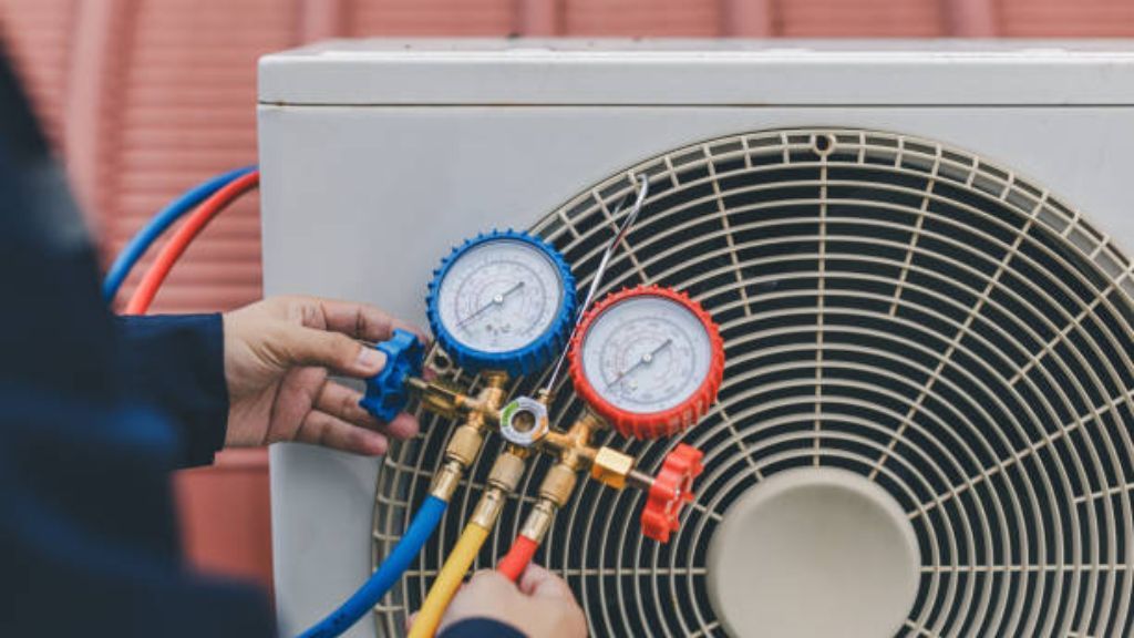 A technician uses a gauge manifold set with blue, yellow, and red hoses to service an outdoor air conditioning unit.