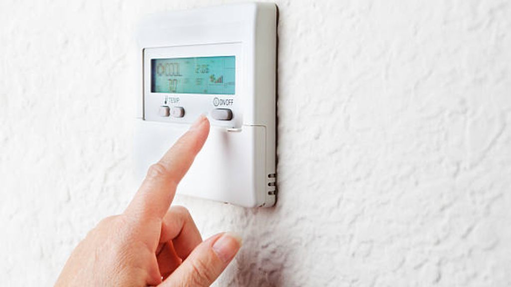 A finger presses a button on a white wall-mounted thermostat to adjust the temperature settings.