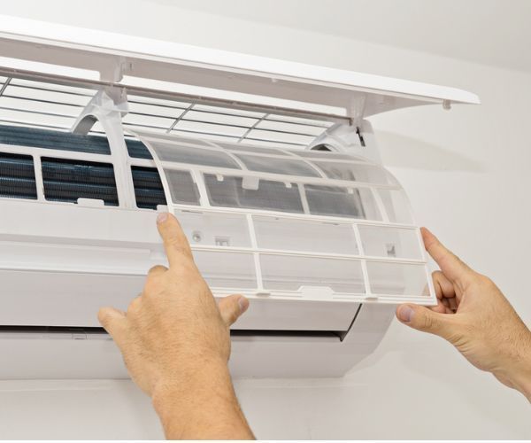 Hands removing a dust filter from an indoor wall-mounted air conditioning unit for cleaning.