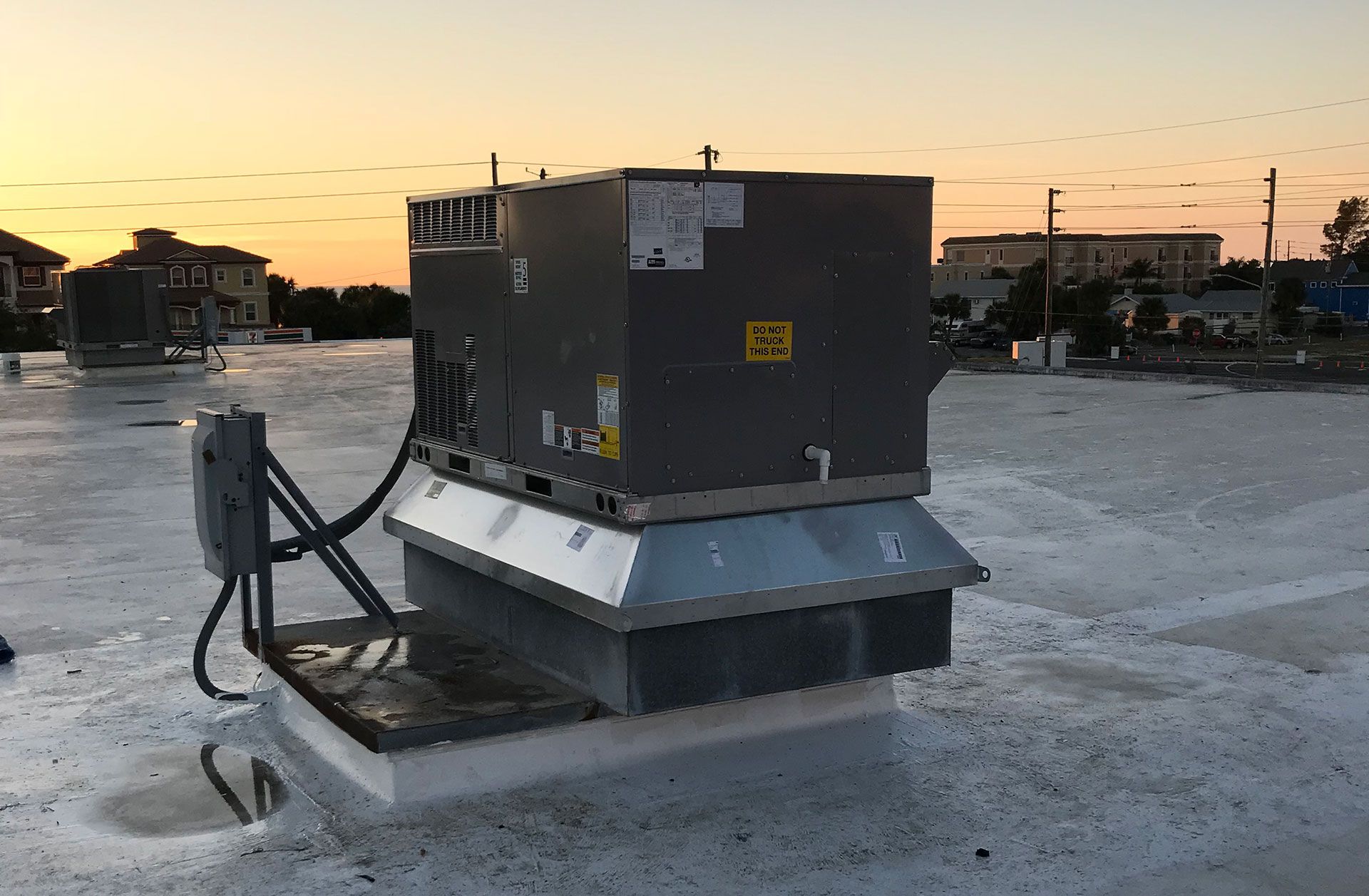 A gray HVAC rooftop unit sits on a flat, white roof during a sunset, with a utility disconnect box attached to its side.