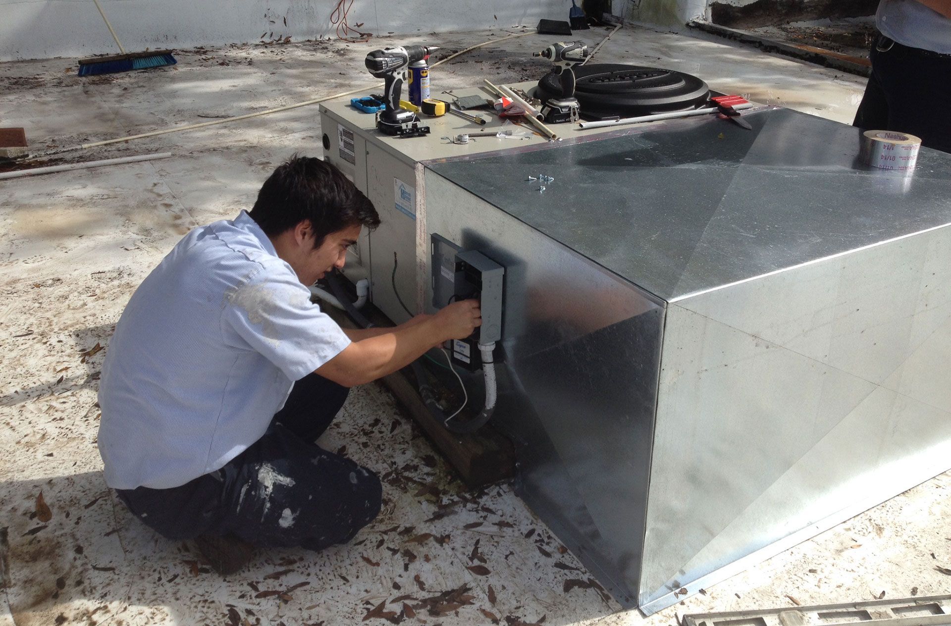 A person wearing a light blue shirt works on electrical wiring on a metal HVAC unit on a flat, debris-covered roof.