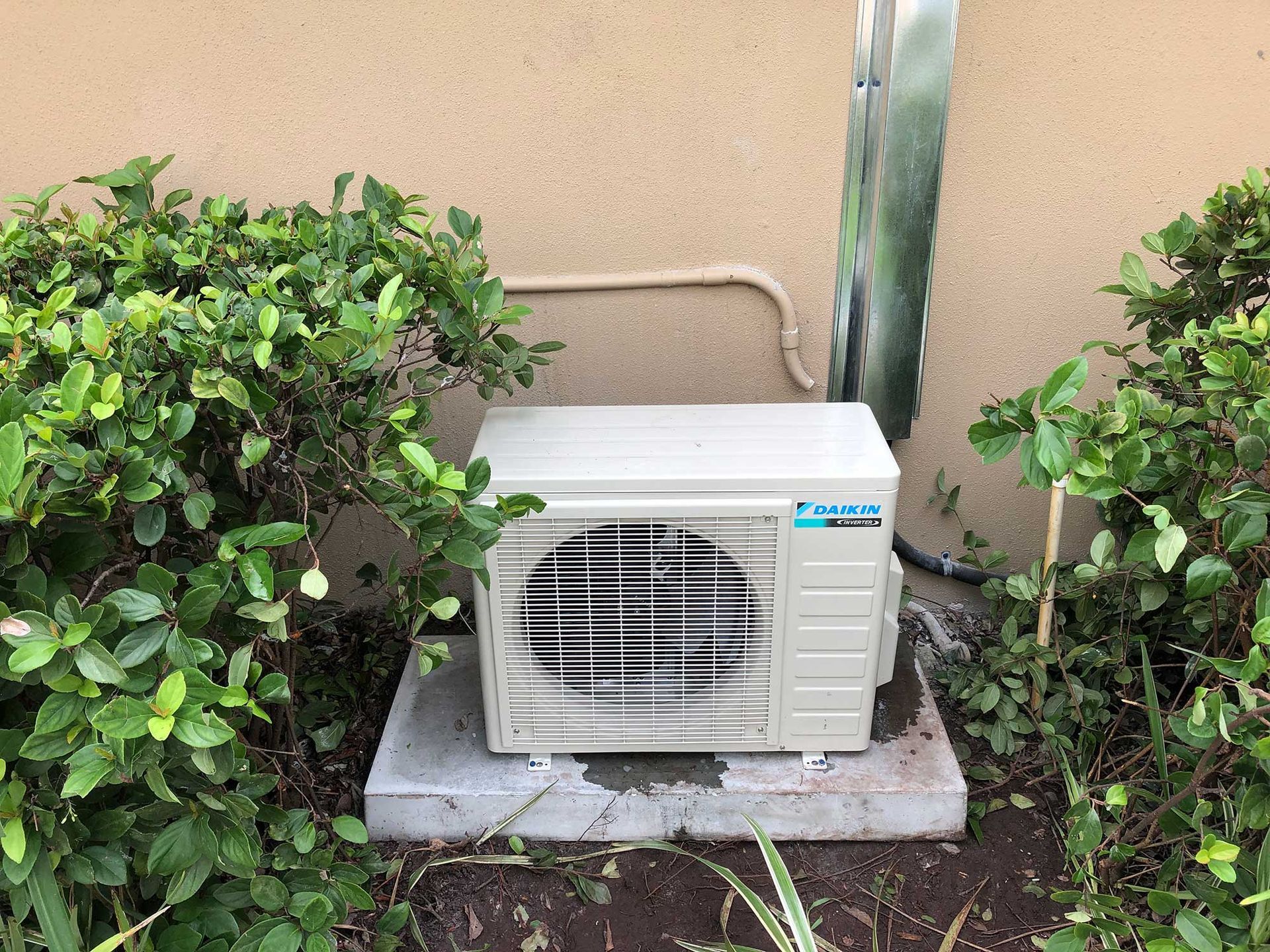 A beige, rectangular outdoor air conditioning condenser unit sits on a concrete pad surrounded by green bushes.
