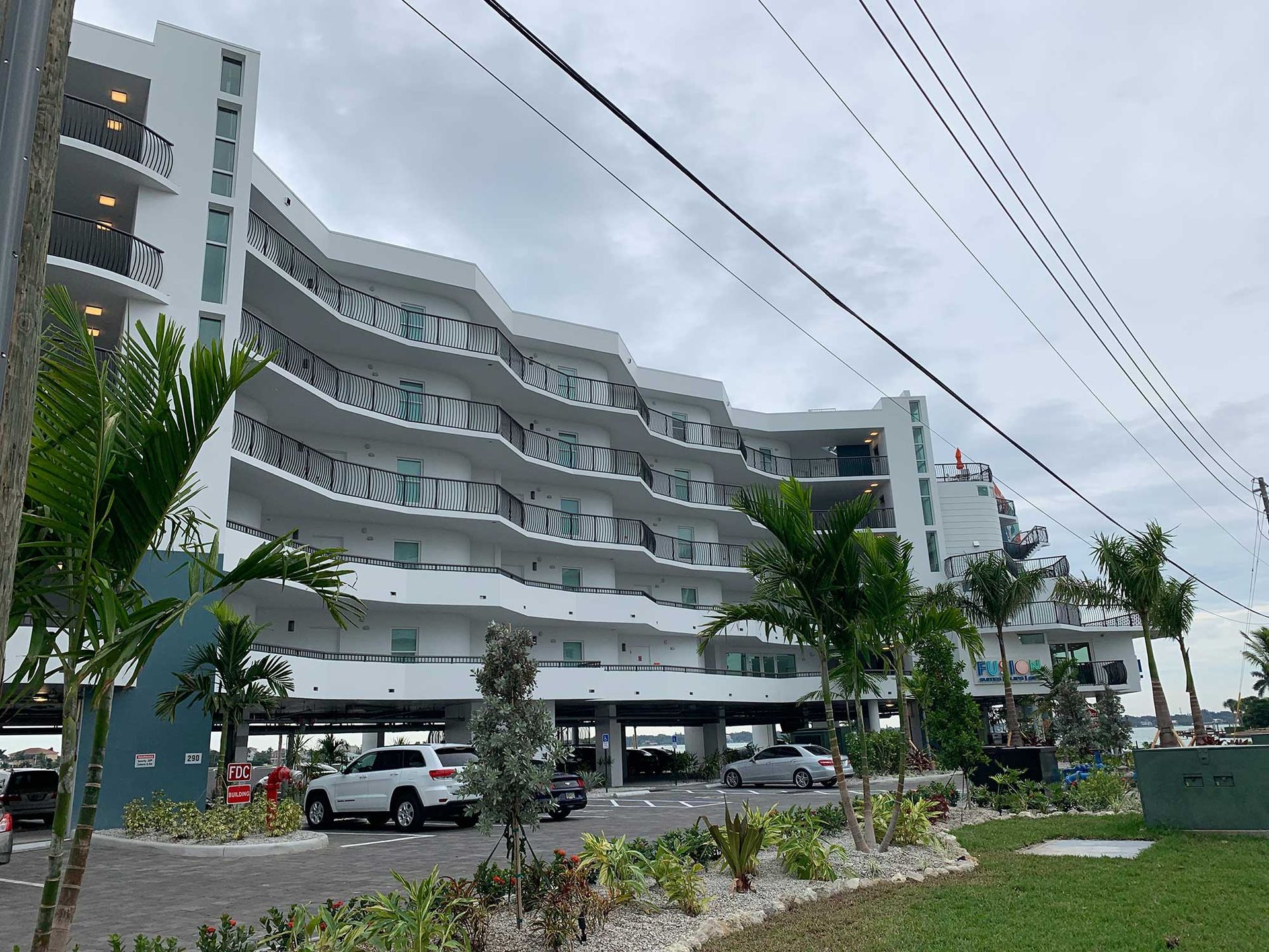 A white multi-story hotel building with a zig-zag facade, balconies, and a parking area at its base under a cloudy sky.