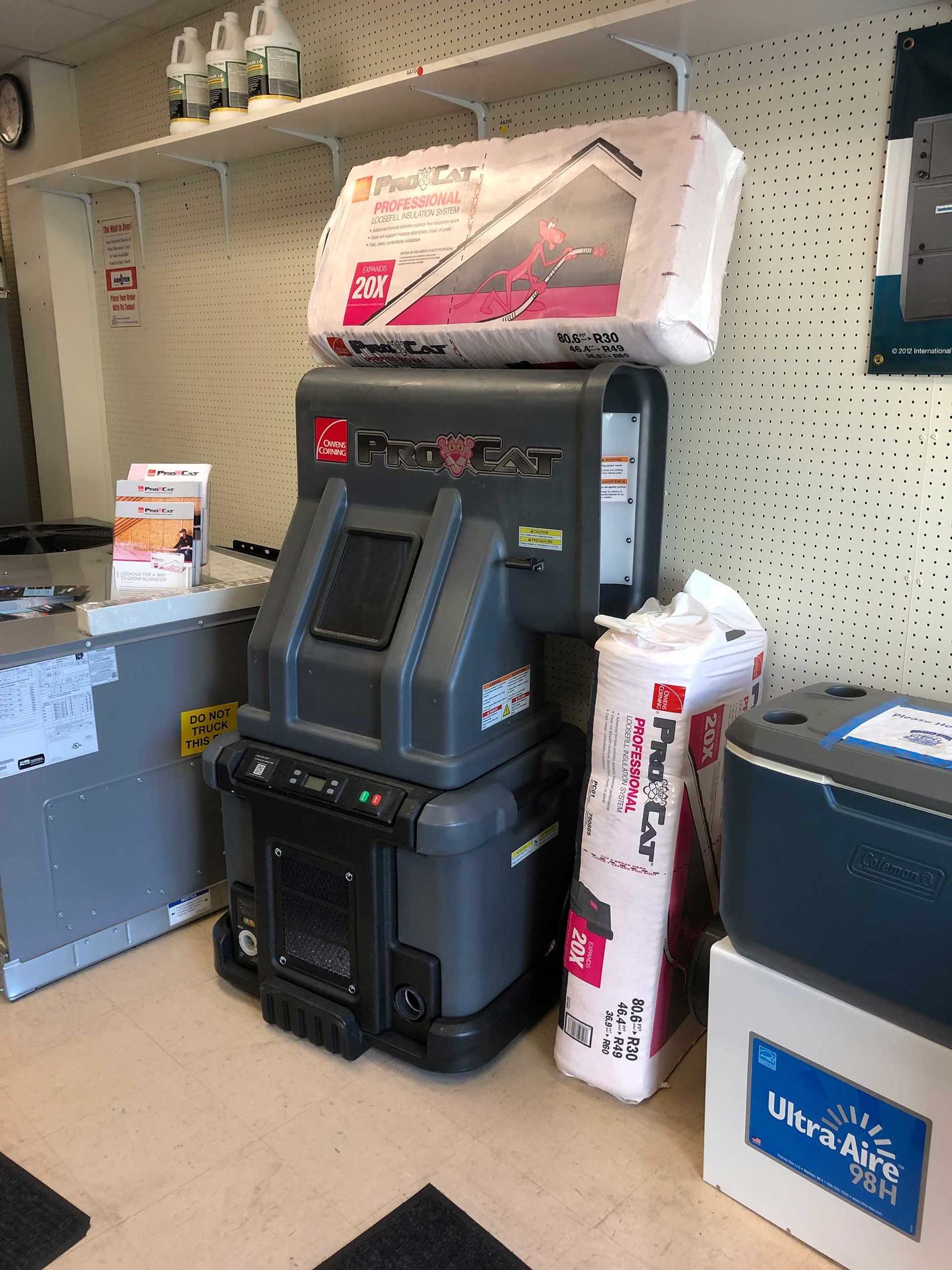 A grey attic insulation blowing machine sits in a retail store with bags of insulation stacked on top and beside it.