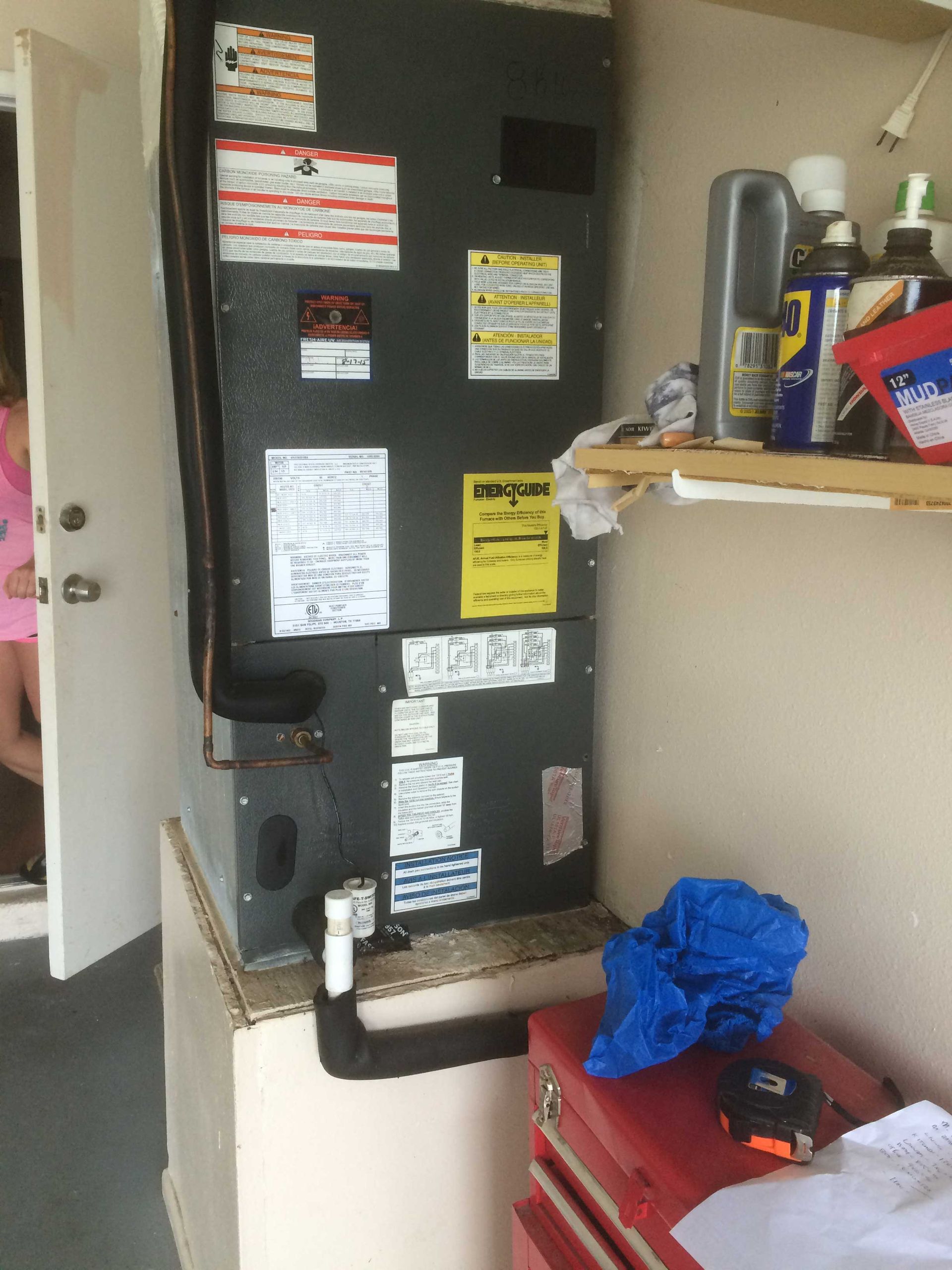A gray HVAC air handler unit stands in a utility closet with multiple instructional labels and a drain line at its base.