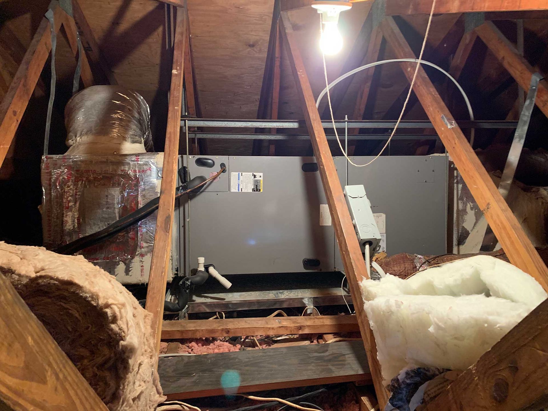 An HVAC air handler unit installed in an unfinished wooden attic, with exposed insulation and a lightbulb hanging above.