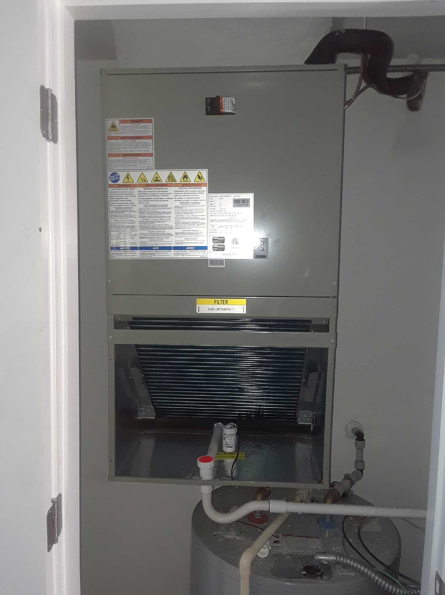 An air handler unit mounted above a water heater in a utility closet, with an exposed cooling coil and drainage pipes.