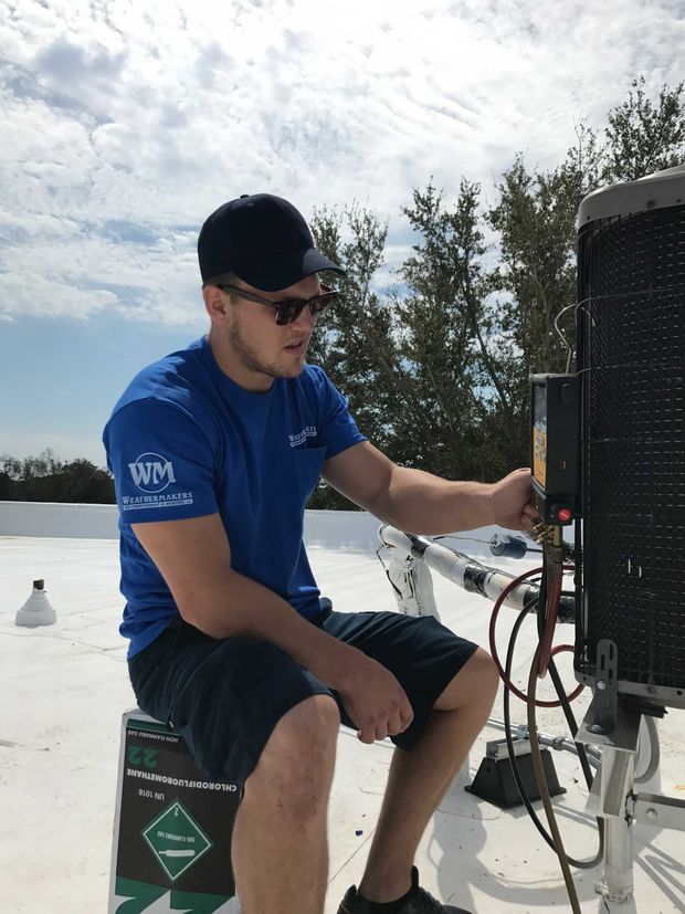 A person in a blue shirt and cap sits on a roof, working on an industrial HVAC unit with tools.