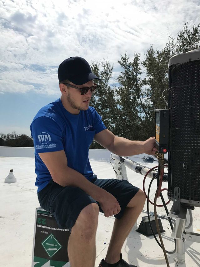 A person in a blue shirt and cap sits on a roof, working on an industrial HVAC unit with tools.