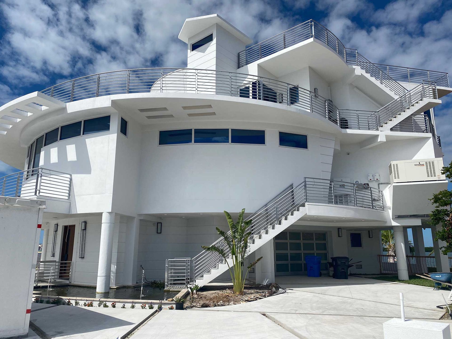 A multi-level, circular white modern house with metal railings, an exterior staircase, and a ground-floor garage.