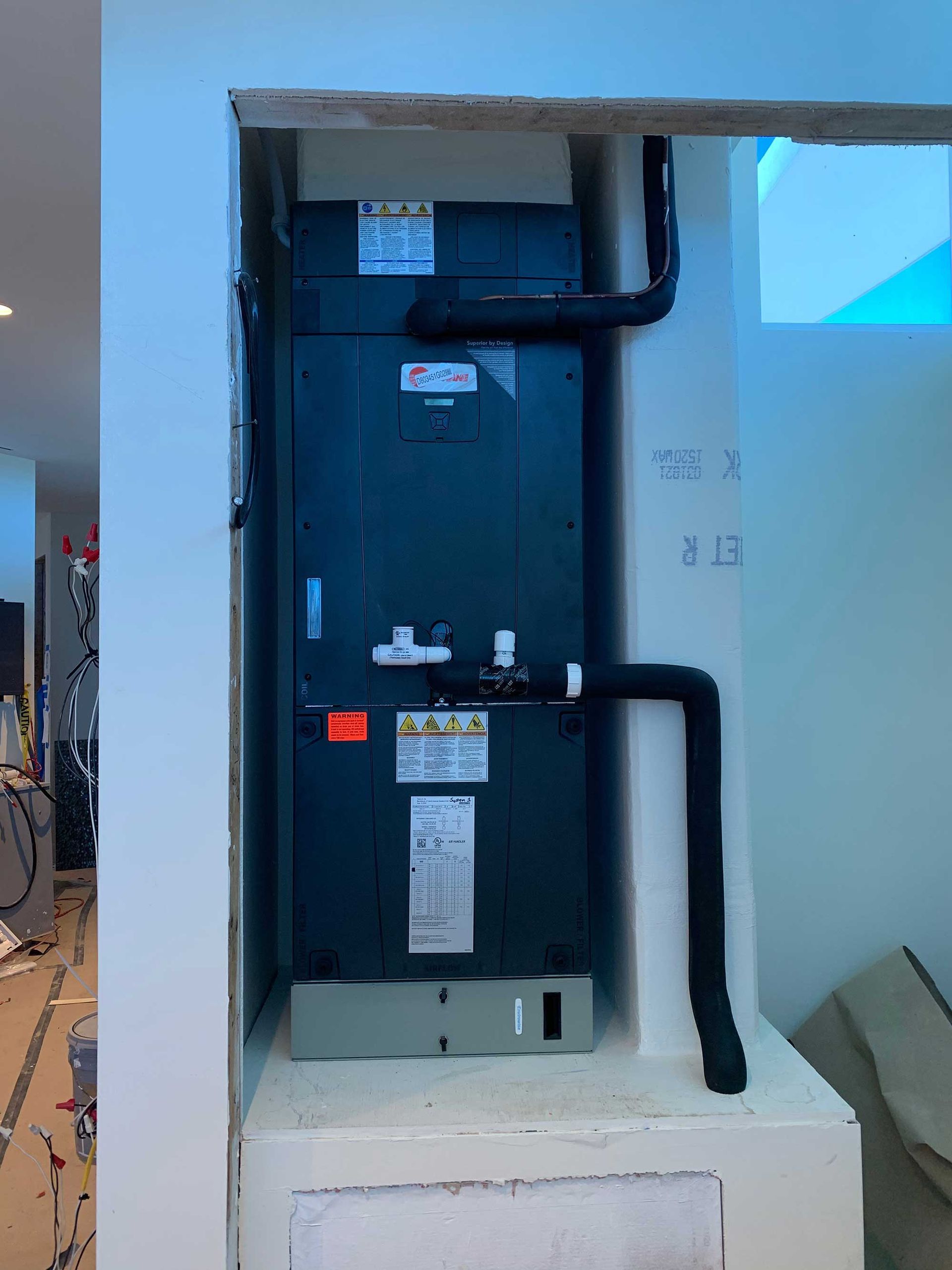 A tall, vertical HVAC air handler unit installed in a wall alcove with black insulated pipes and white drain lines.