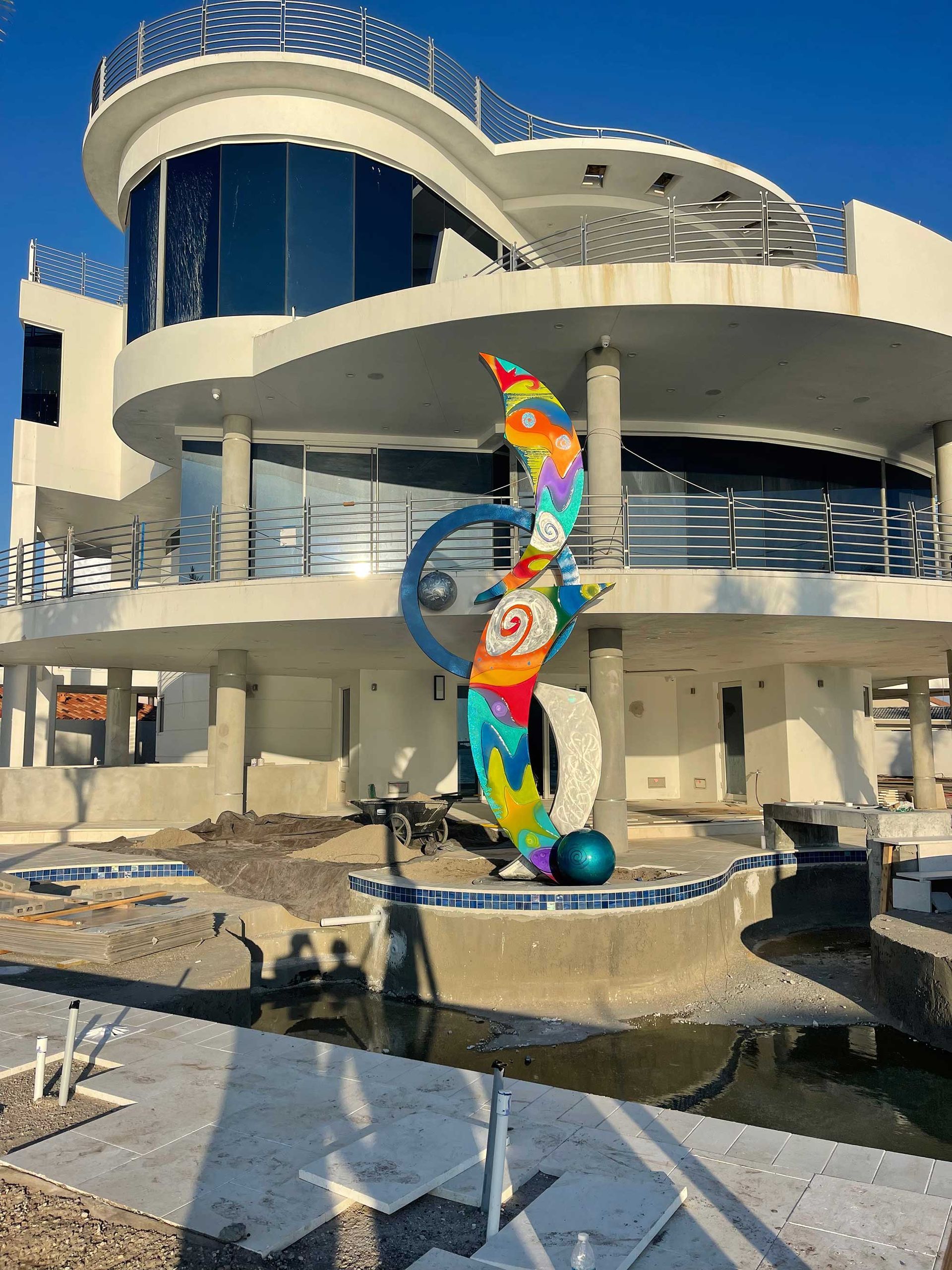 A multi-colored abstract sculpture stands in front of a modern, curved house with tinted windows under a clear blue sky.
