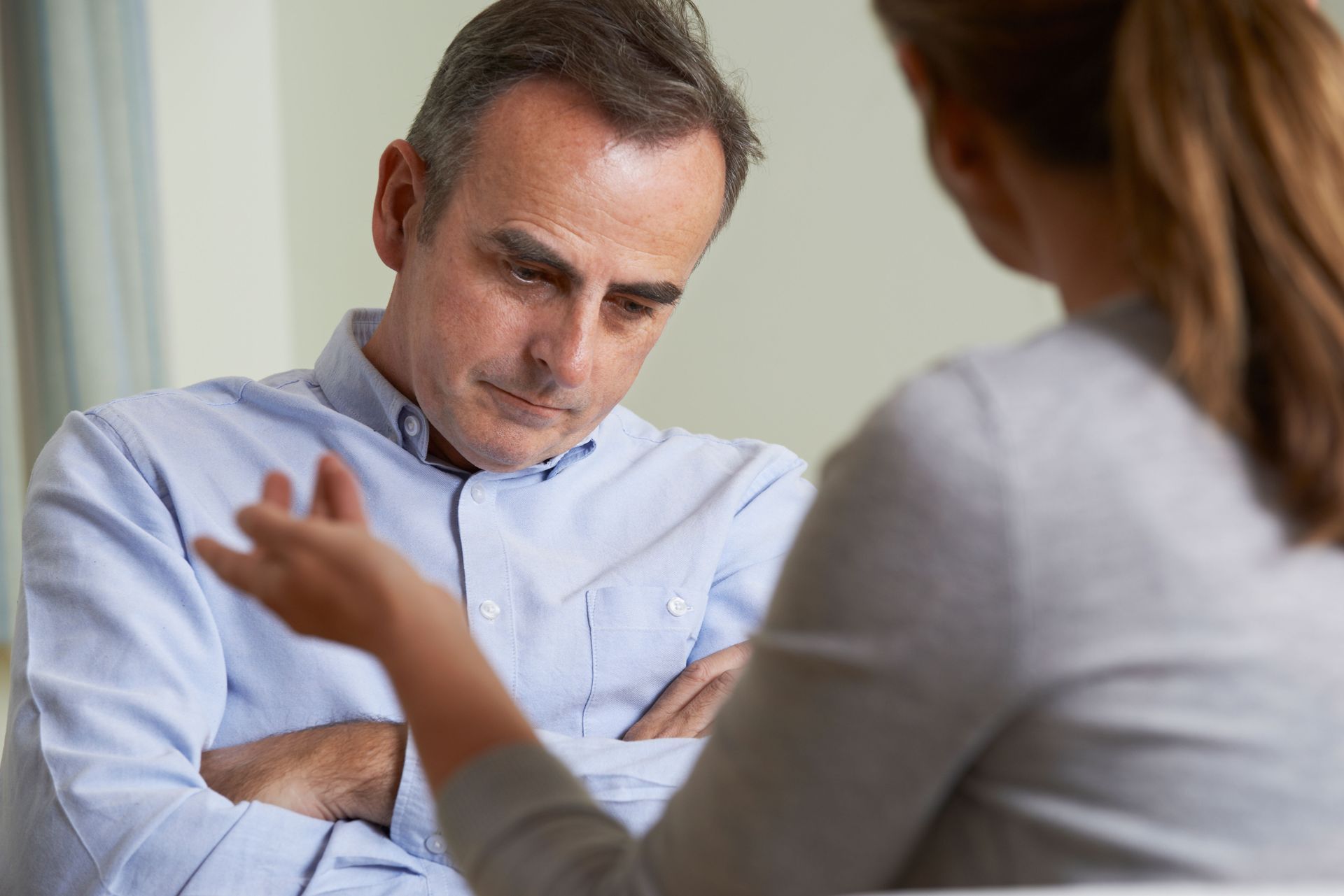 A person in a light blue shirt listens with crossed arms as another person speaks to them in a soft, lit interior setting.