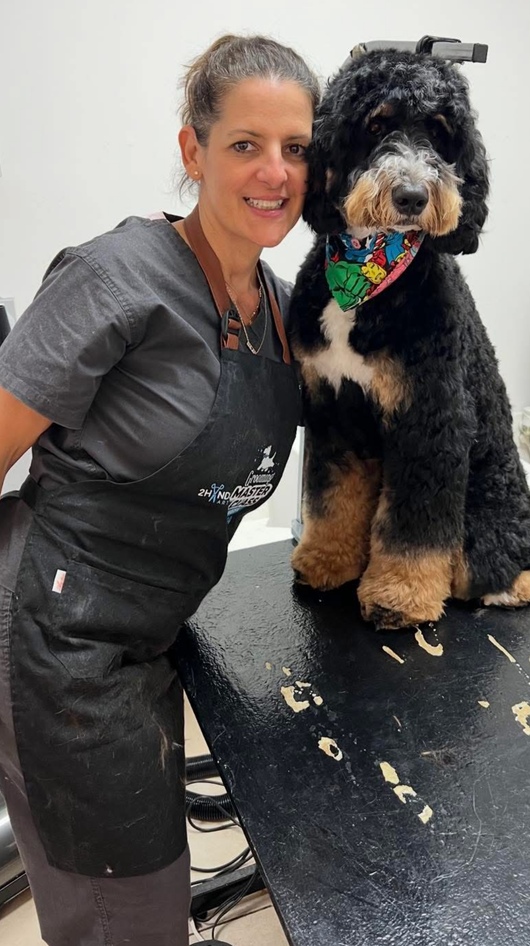 Woman in gray uniform standing beside a black and tan dog on a grooming table, both looking at the camera.