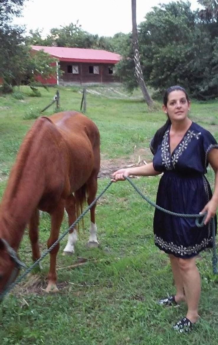 A woman in a blue dress holds a horse's lead rope on a grassy field, with a red-roofed building in the background.