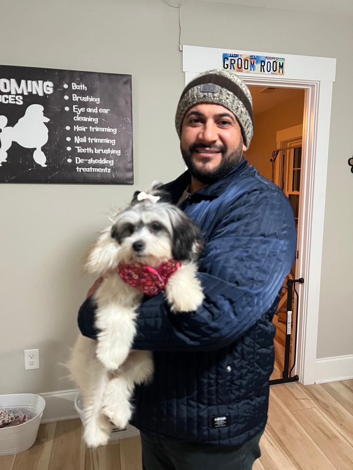 A man in a blue jacket holding a small, fluffy dog with a red scarf in a grooming shop.