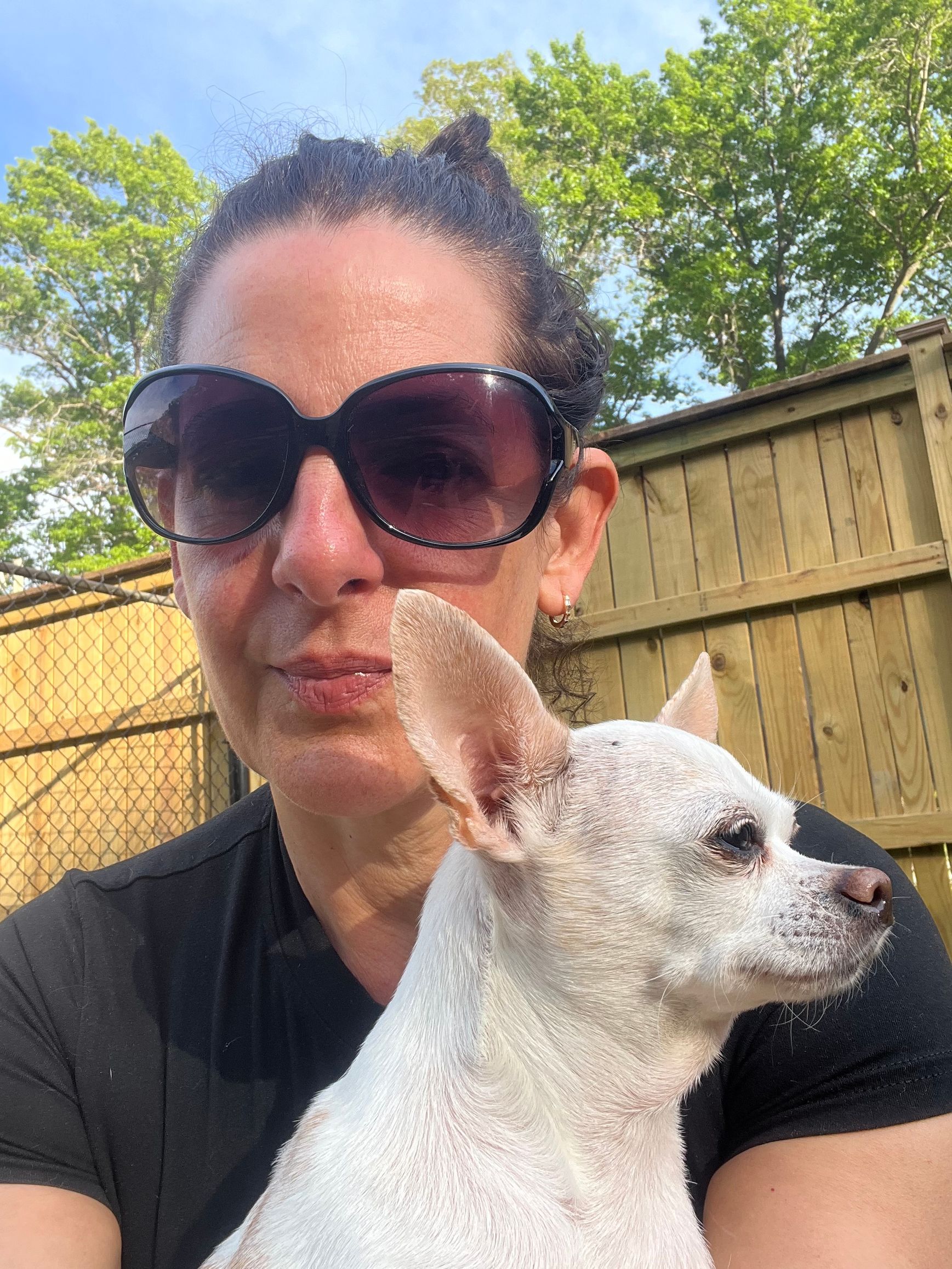 Woman in sunglasses holding a small white dog outdoors by a wooden fence.