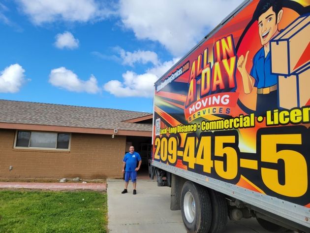 A person in a blue uniform stands by a brightly colored "All In A Day Moving Services" truck parked at a residential home.