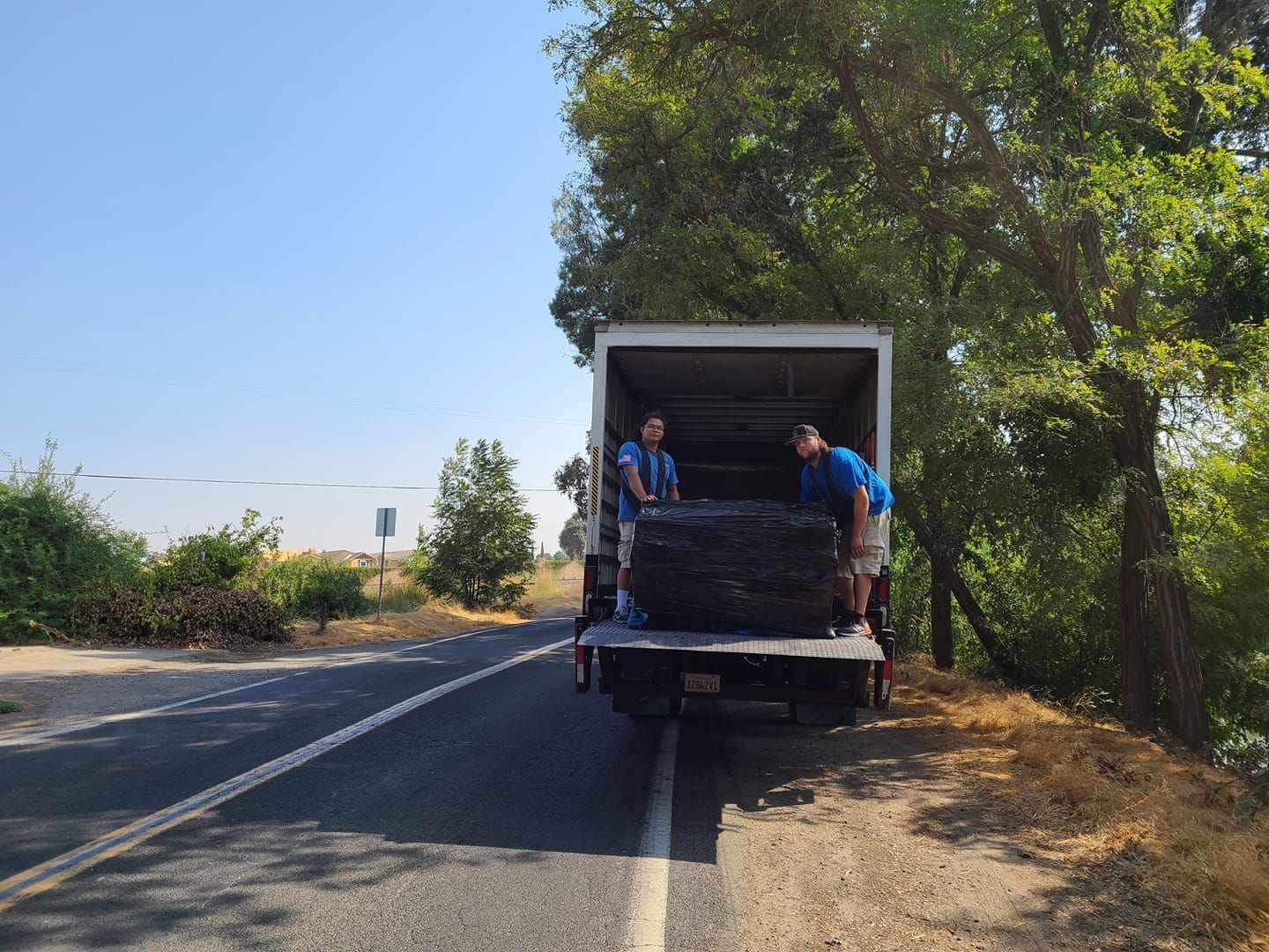 Two people in blue shirts load a large, dark object into the back of a box truck parked on a roadside near trees.