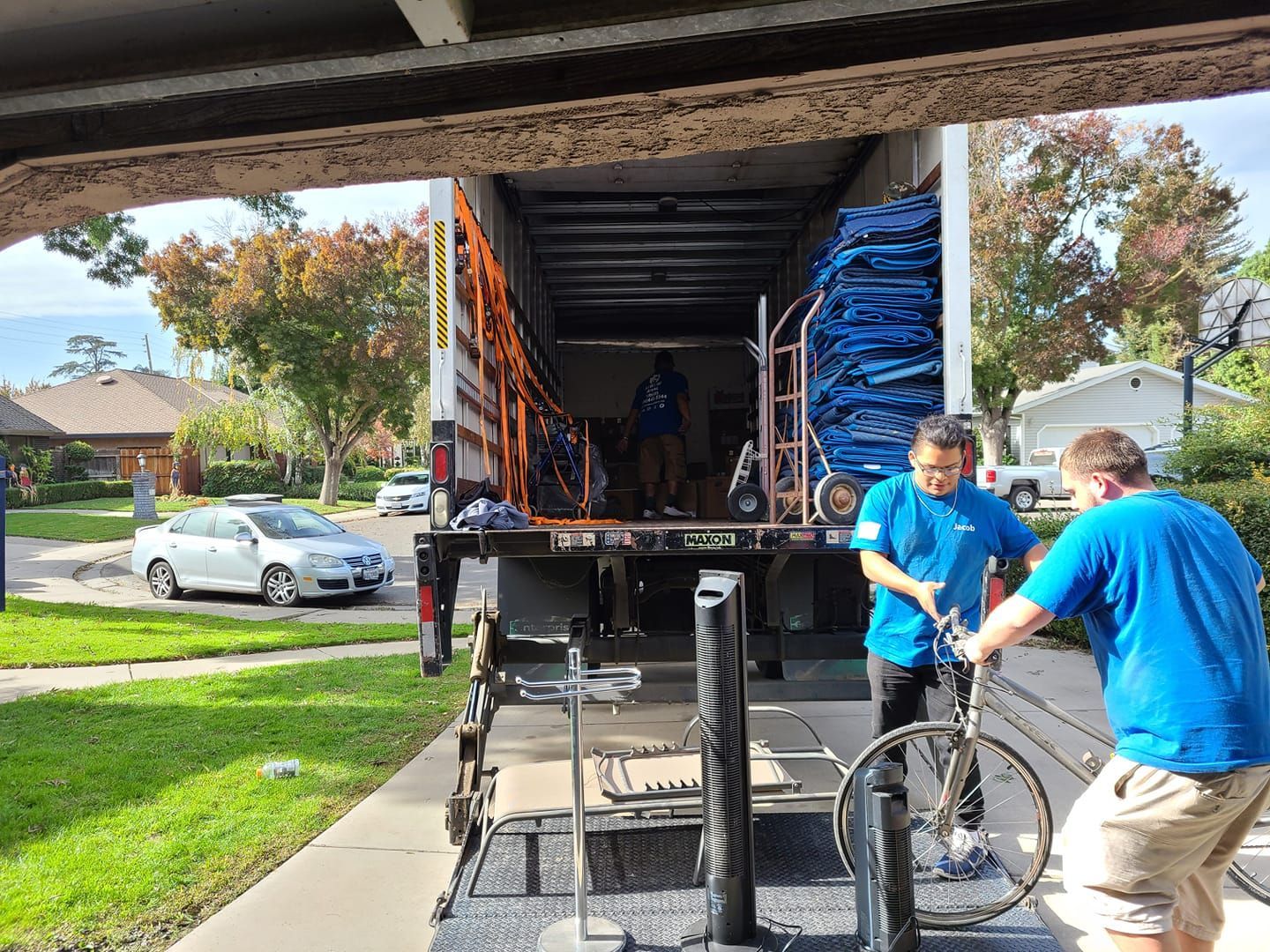 Two people in blue shirts load furniture and a bicycle into the back of a moving truck parked on a residential street.
