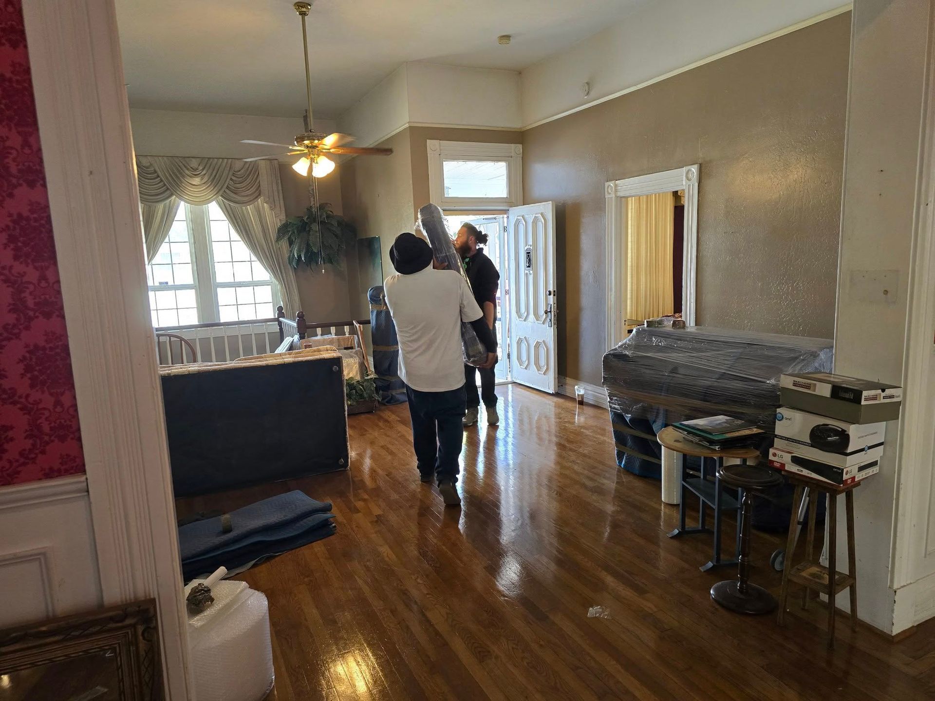 Two people carrying furniture through a bright room with hardwood floors, a large window, and open doorways.