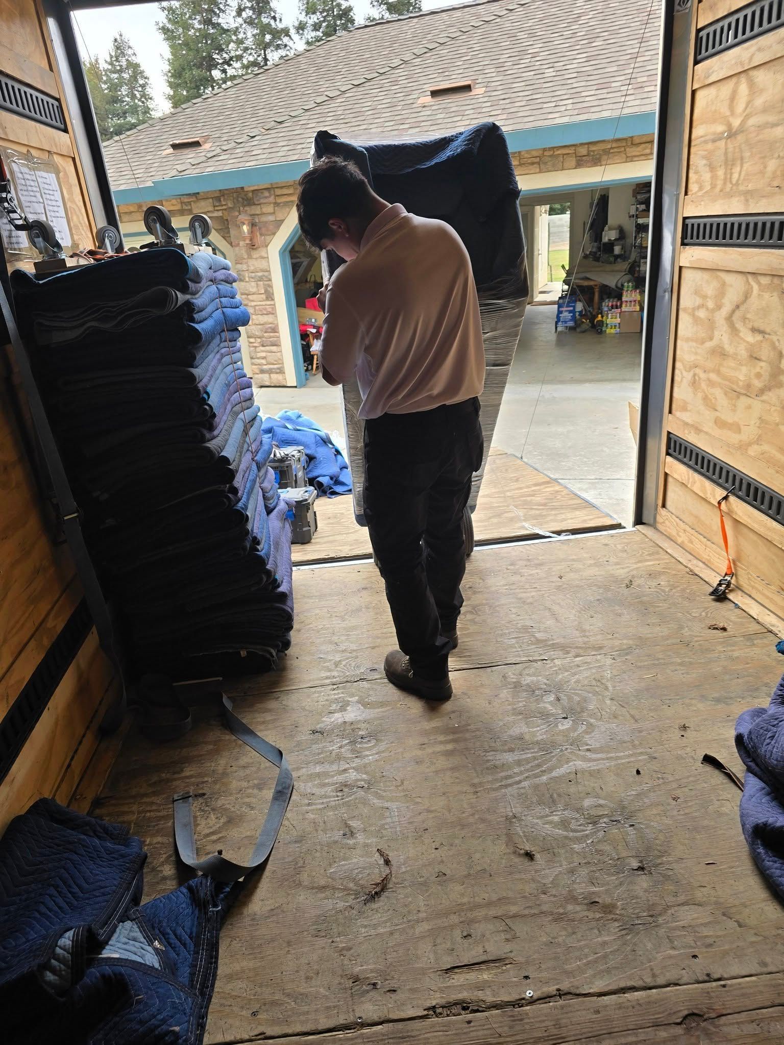 A person in a light shirt and dark pants carries a wrapped item into a moving truck, with stacks of folded pads nearby.