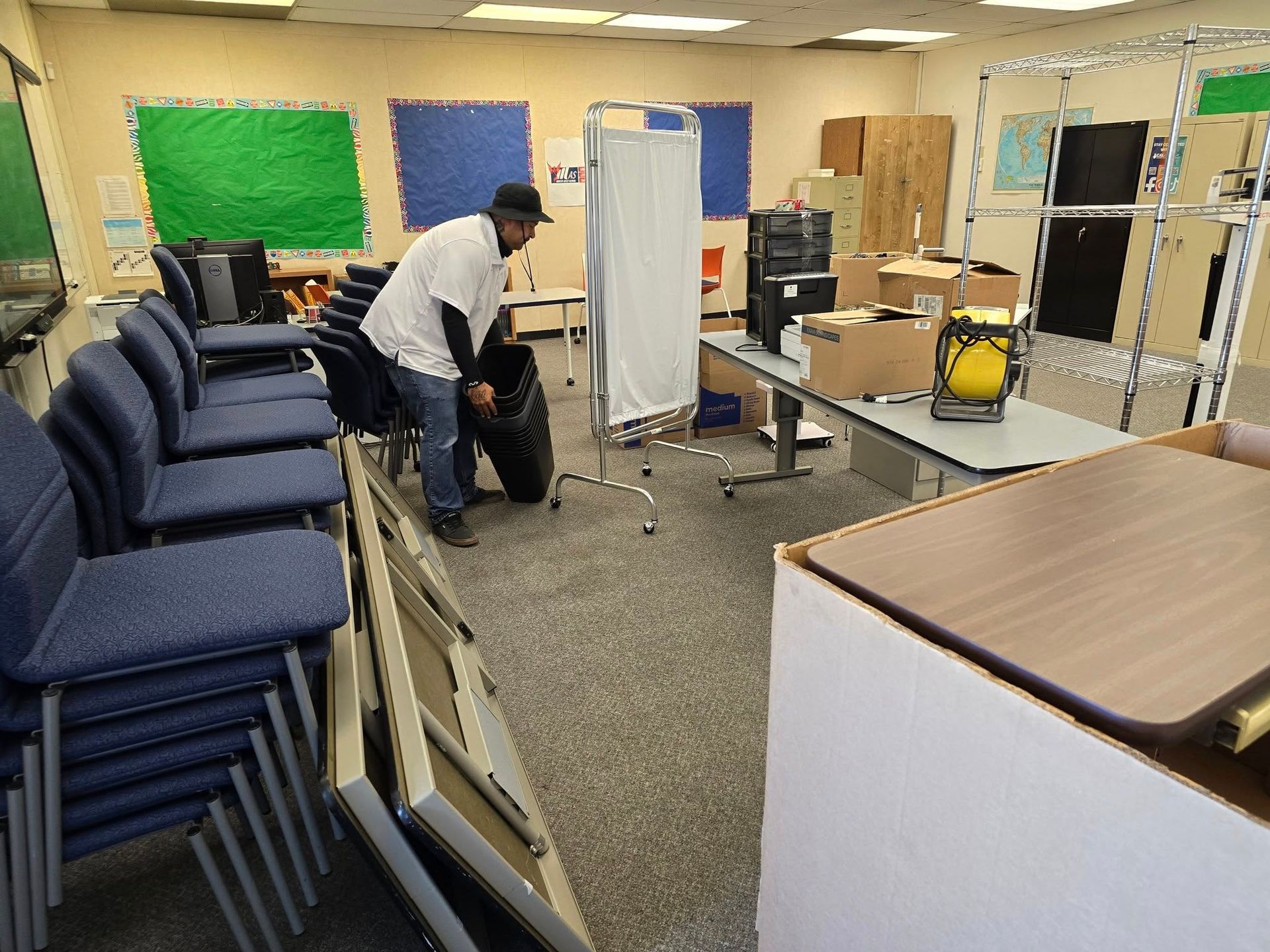 A person moves a black bag in an office or classroom filled with stacks of blue chairs, desks, and office supplies.