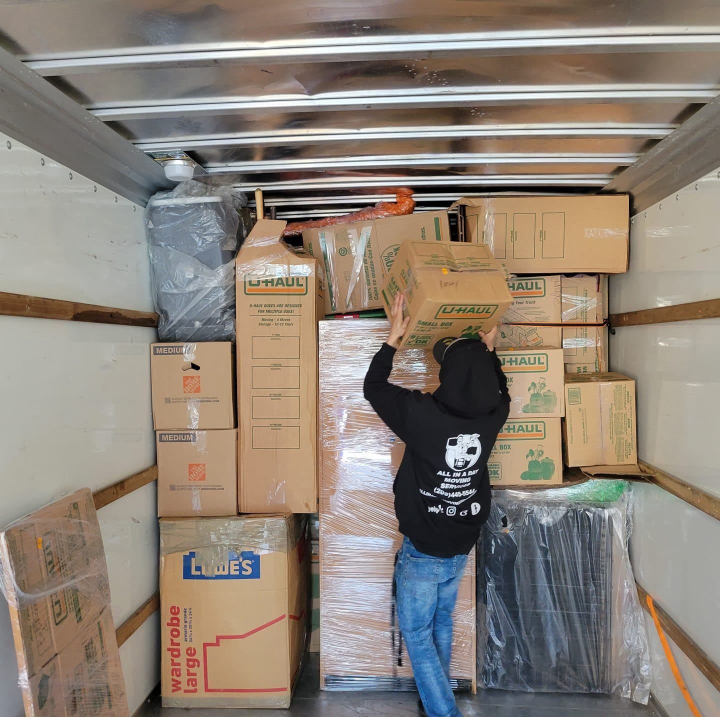 A person wearing a black hoodie and blue jeans stacks a cardboard box inside a moving truck filled with packed boxes.
