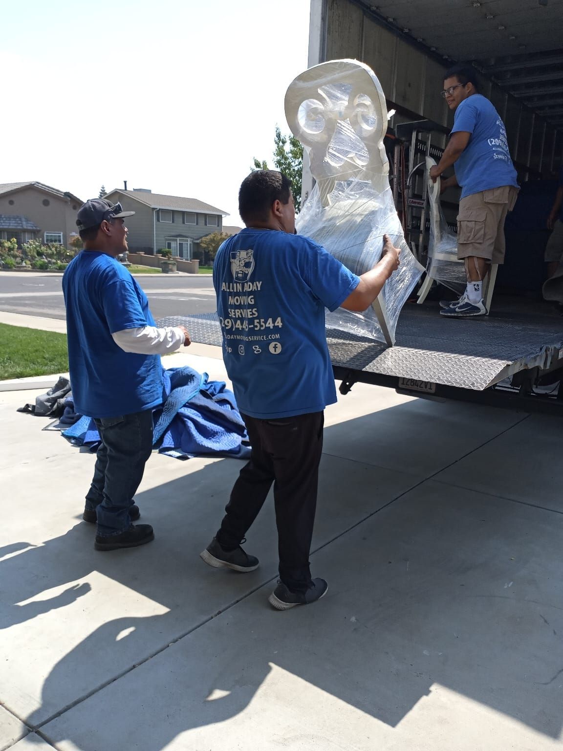 Three movers in blue shirts load a shrink-wrapped chair into a moving truck parked on a residential driveway.