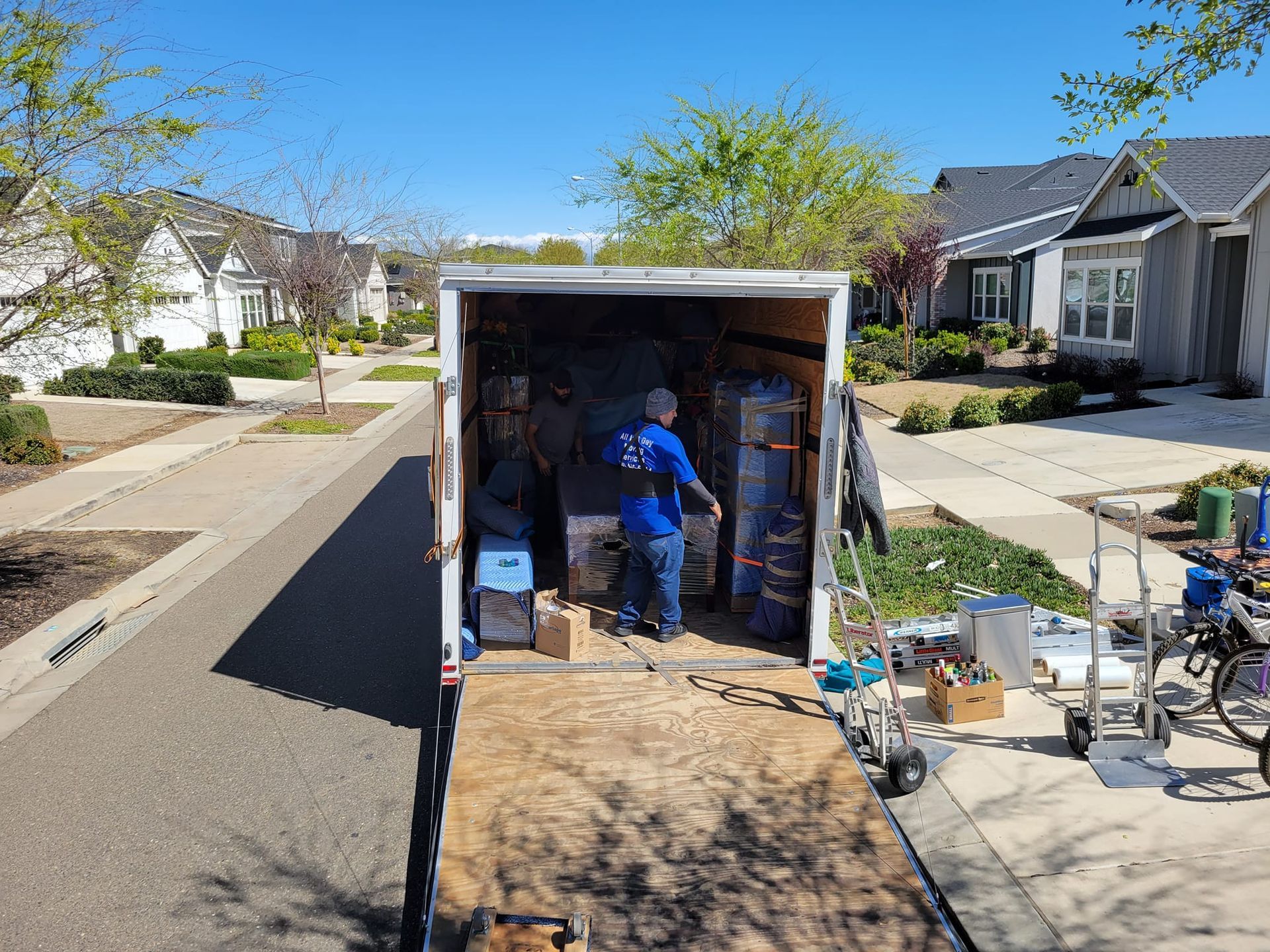 A person in a blue shirt works inside a parked moving truck on a sunny residential street with houses in the background.