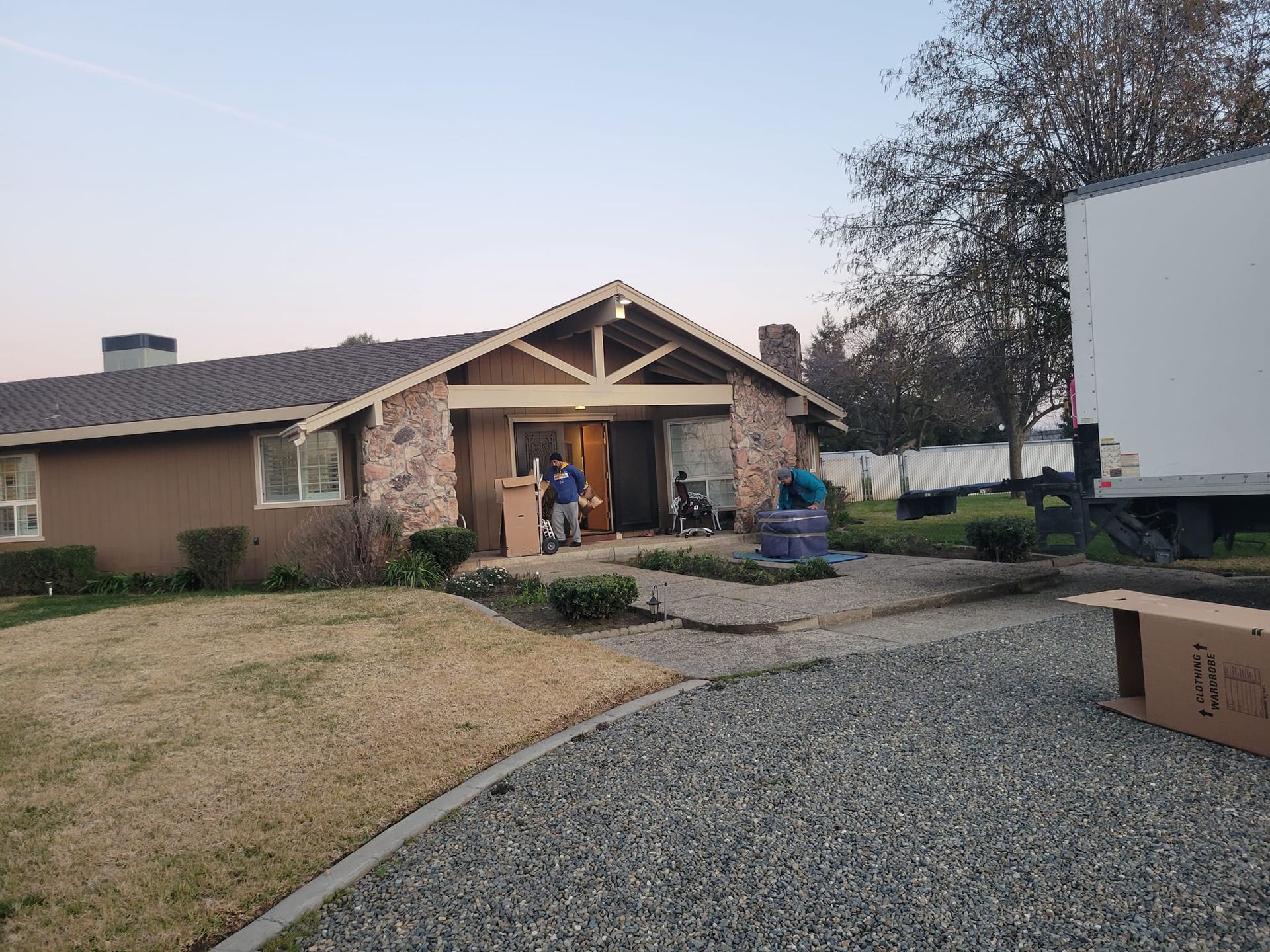 People move items out of a tan house with stone accents into a moving truck in the driveway.
