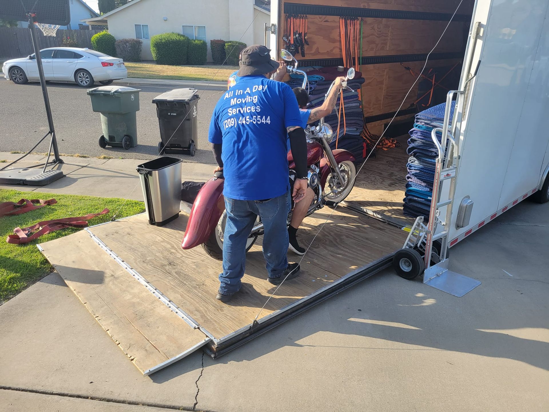 A person in a blue shirt maneuvers a red motorcycle into the back of an open utility trailer on a residential driveway.