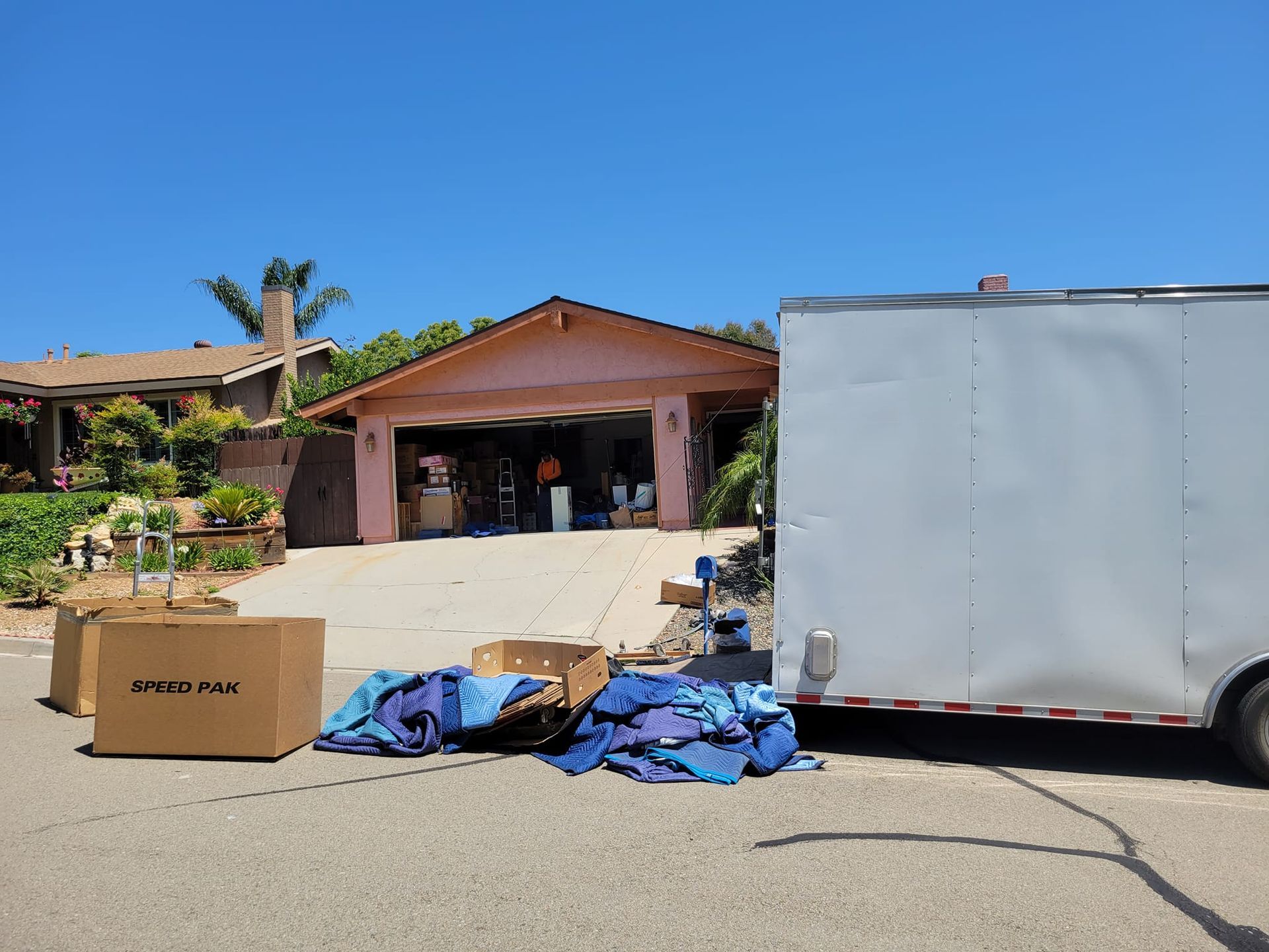 A pink house with an open garage, a white cargo trailer, and moving boxes and blue blankets on a suburban driveway.