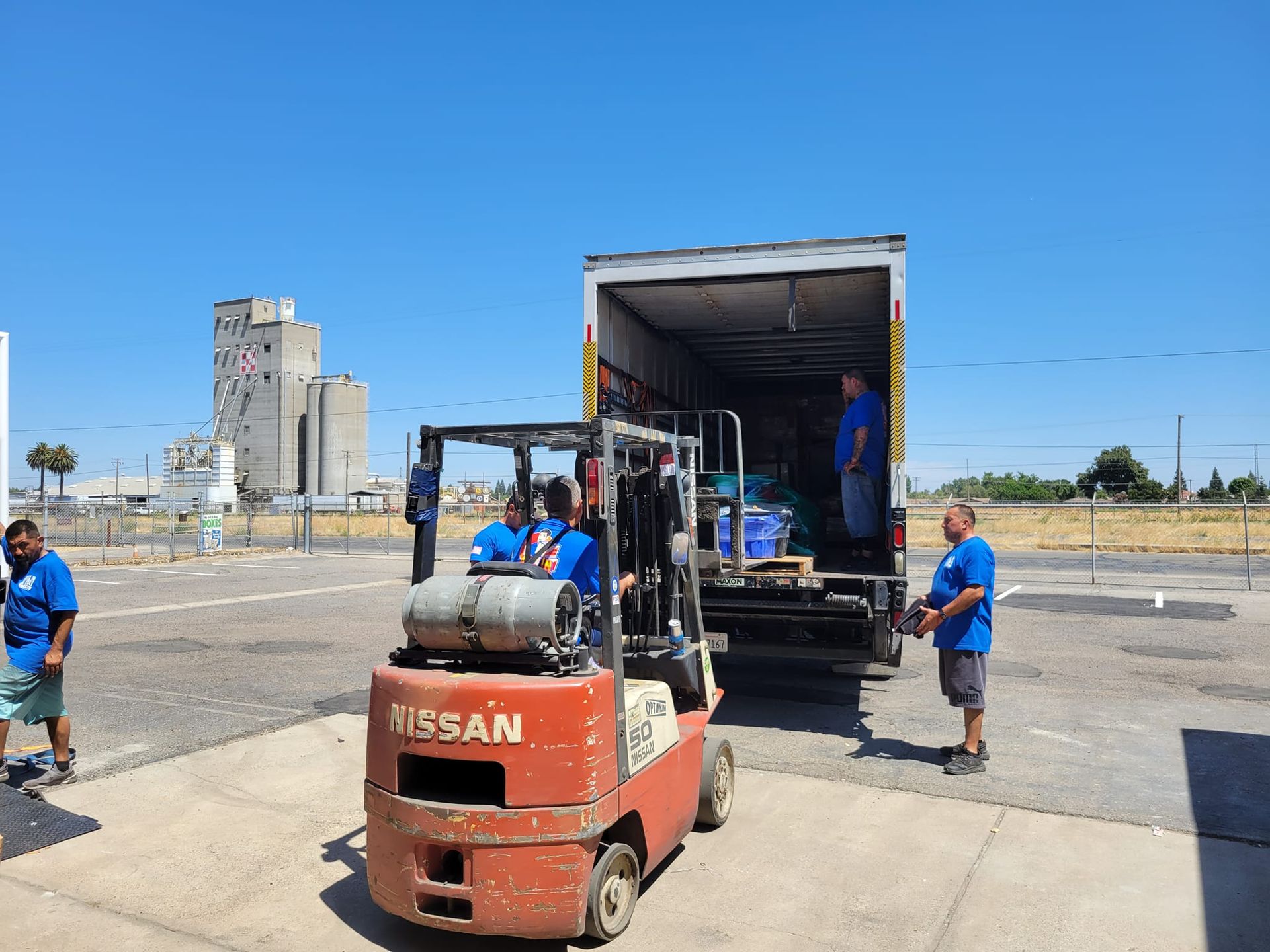 A forklift and a truck are parked in a sunny lot, with workers loading supplies into the open trailer.