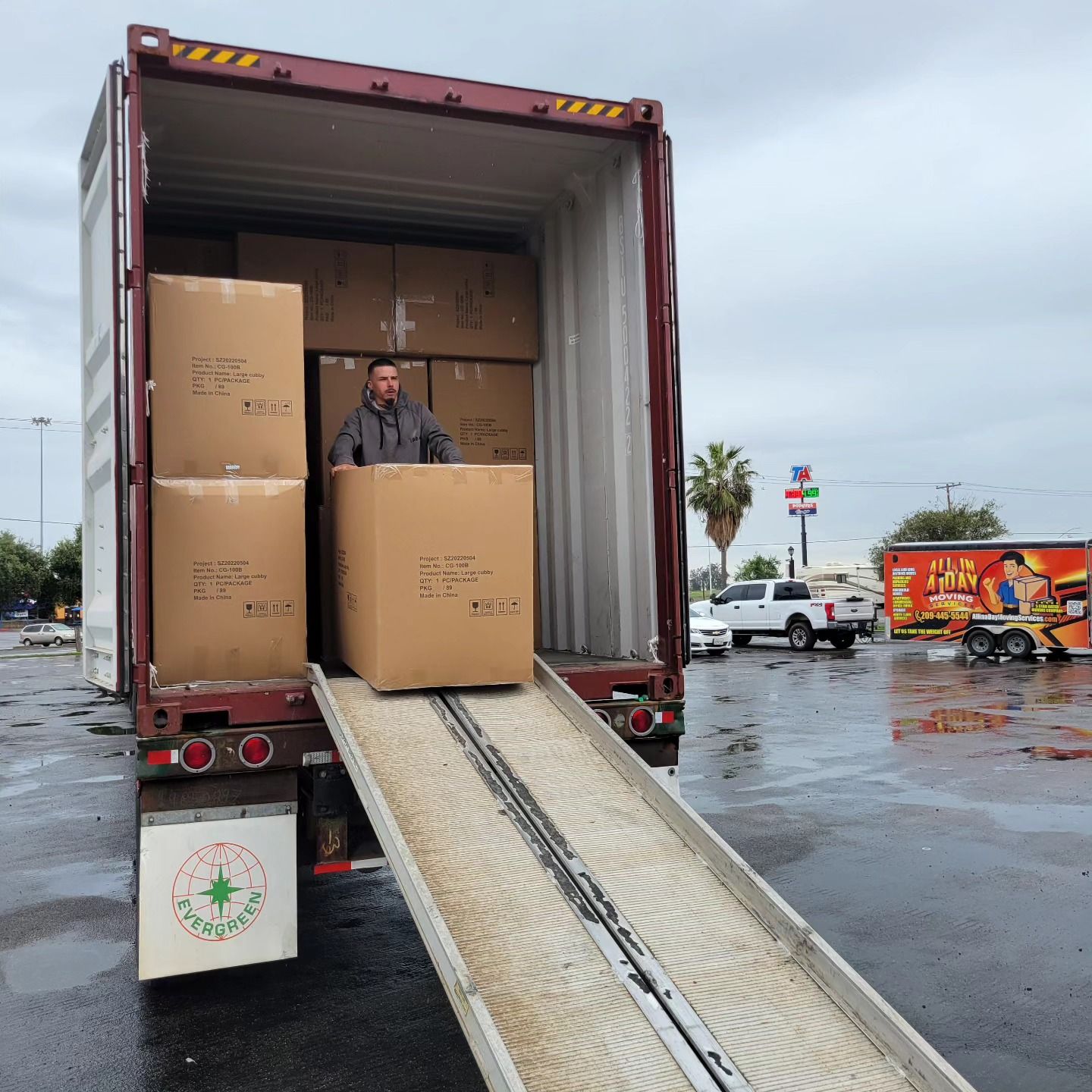 A person moves a large cardboard box down a ramp from inside a shipping container in an outdoor parking lot.