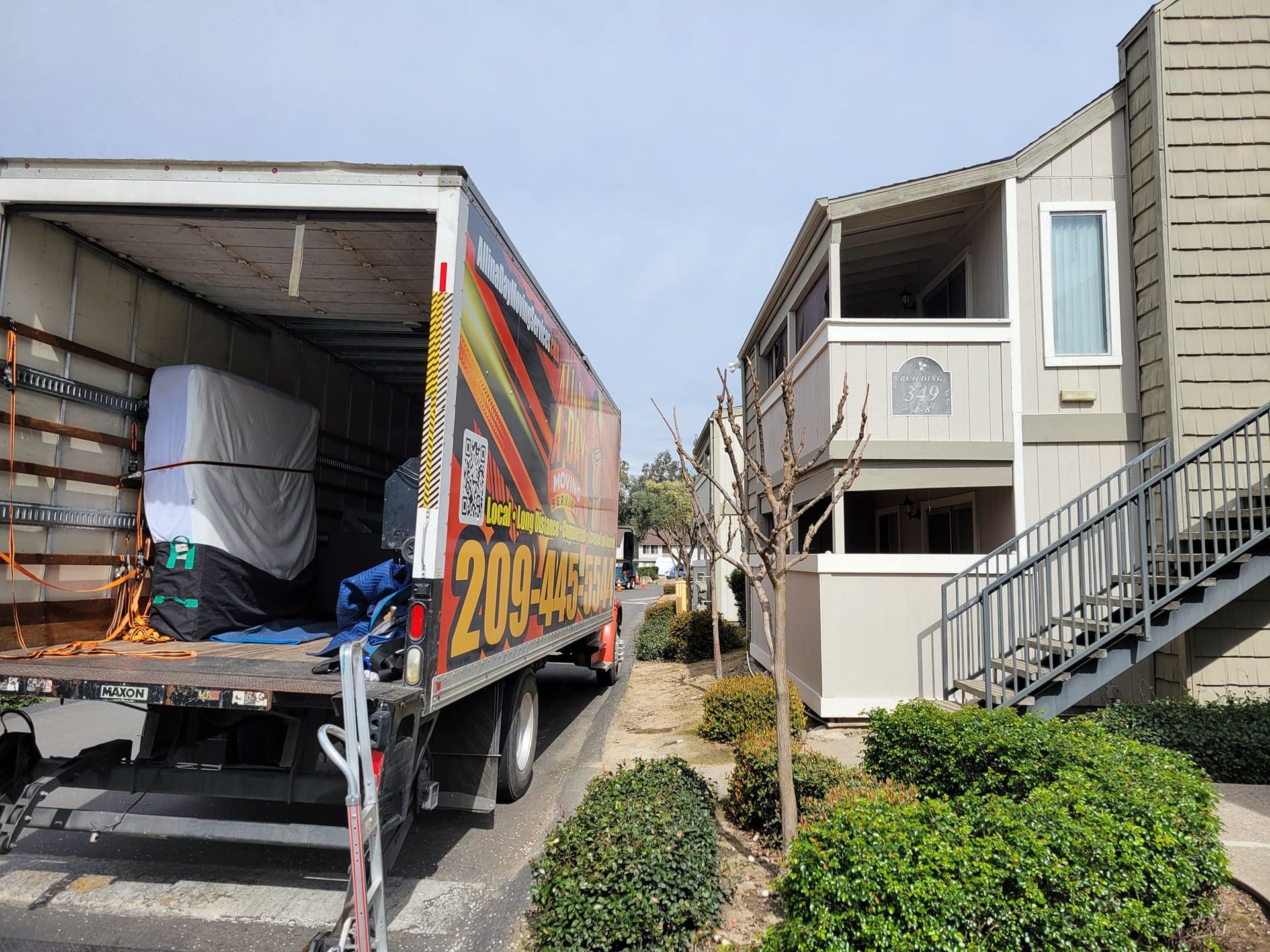 A moving truck with an open back parked next to a multi-story apartment building with outdoor stairs.