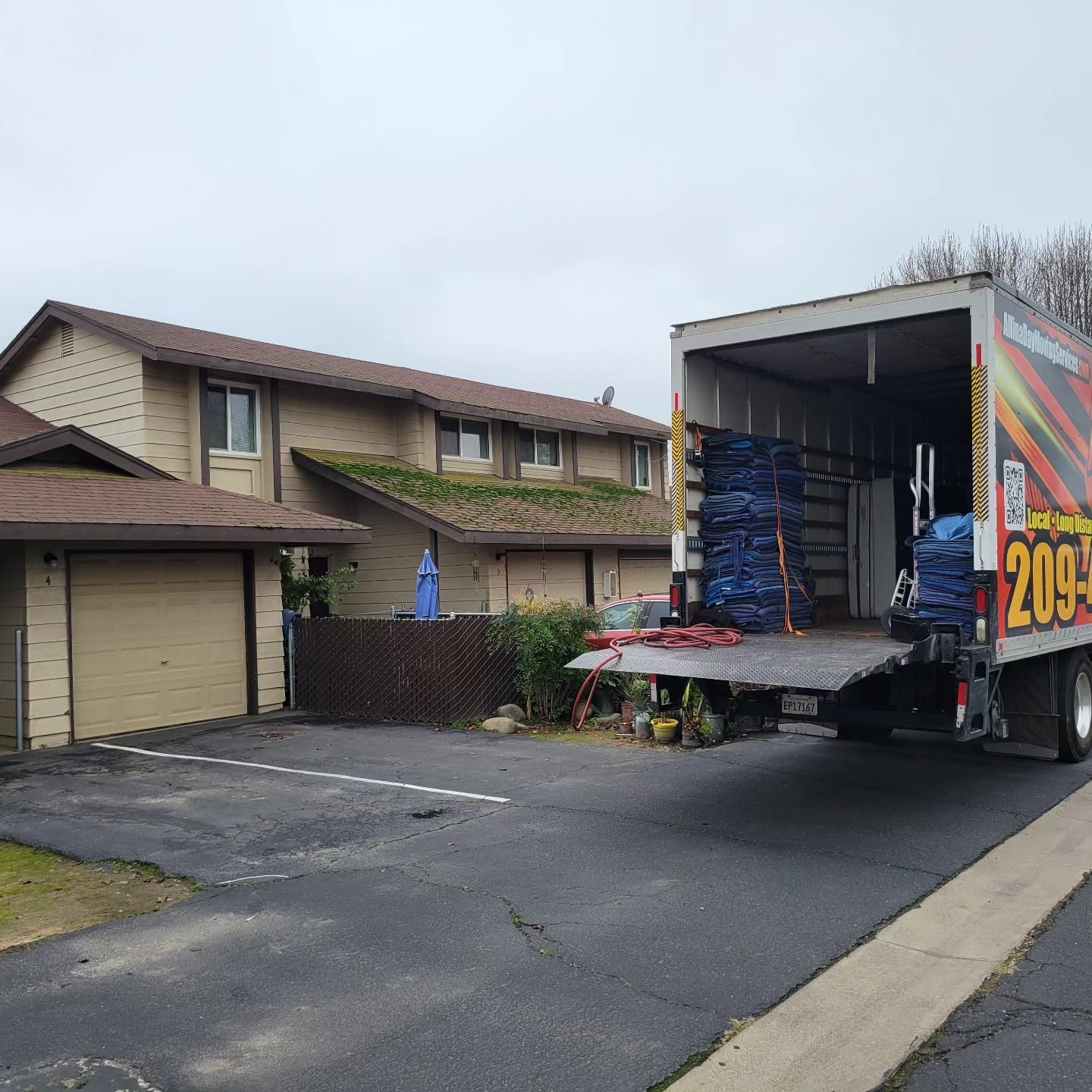 A moving truck parked in the driveway of a tan two-story multi-family house on an overcast day.