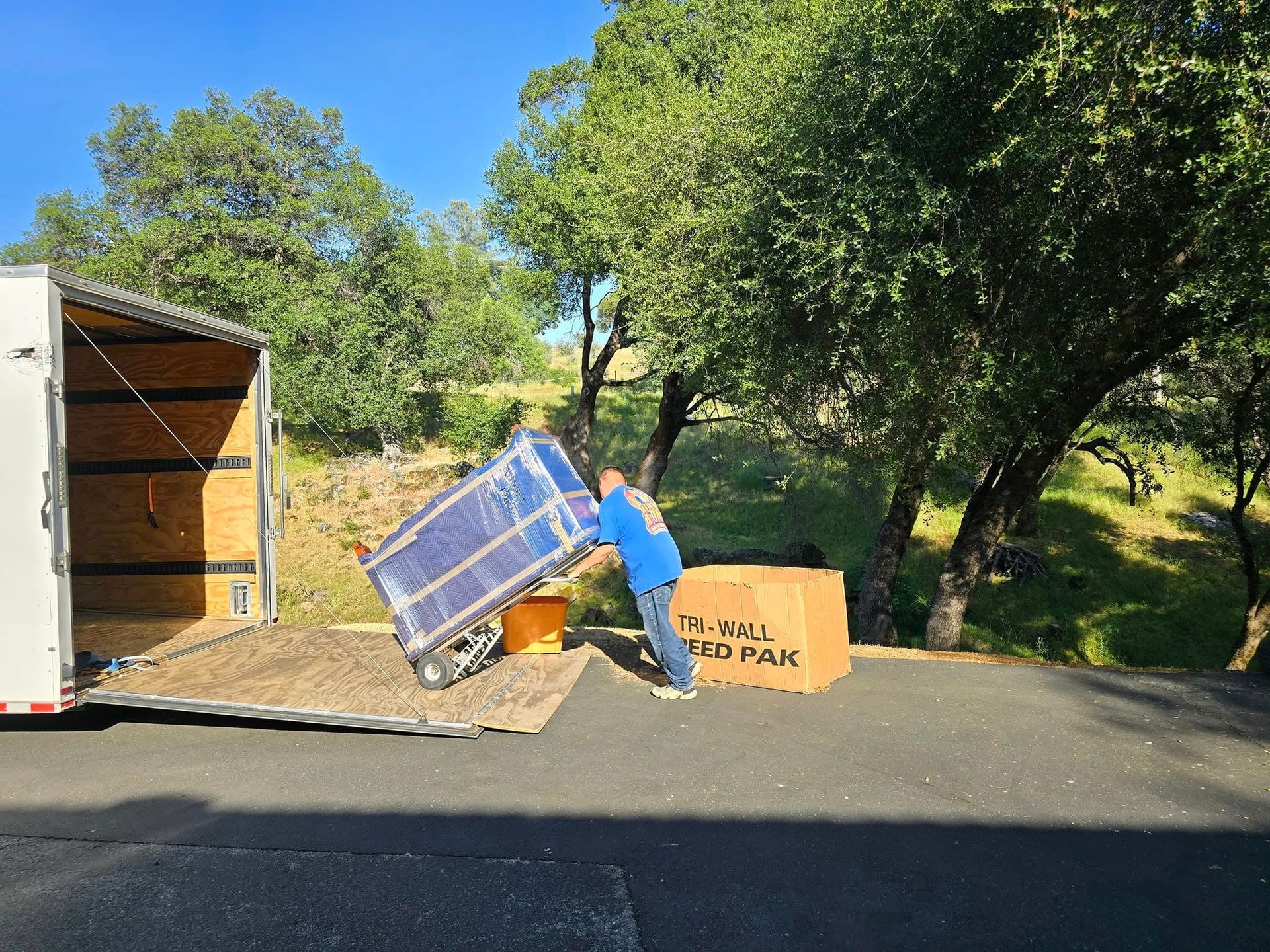A person in a blue shirt maneuvers a large, shrink-wrapped item into the back of a parked trailer on a sunny, rural day.