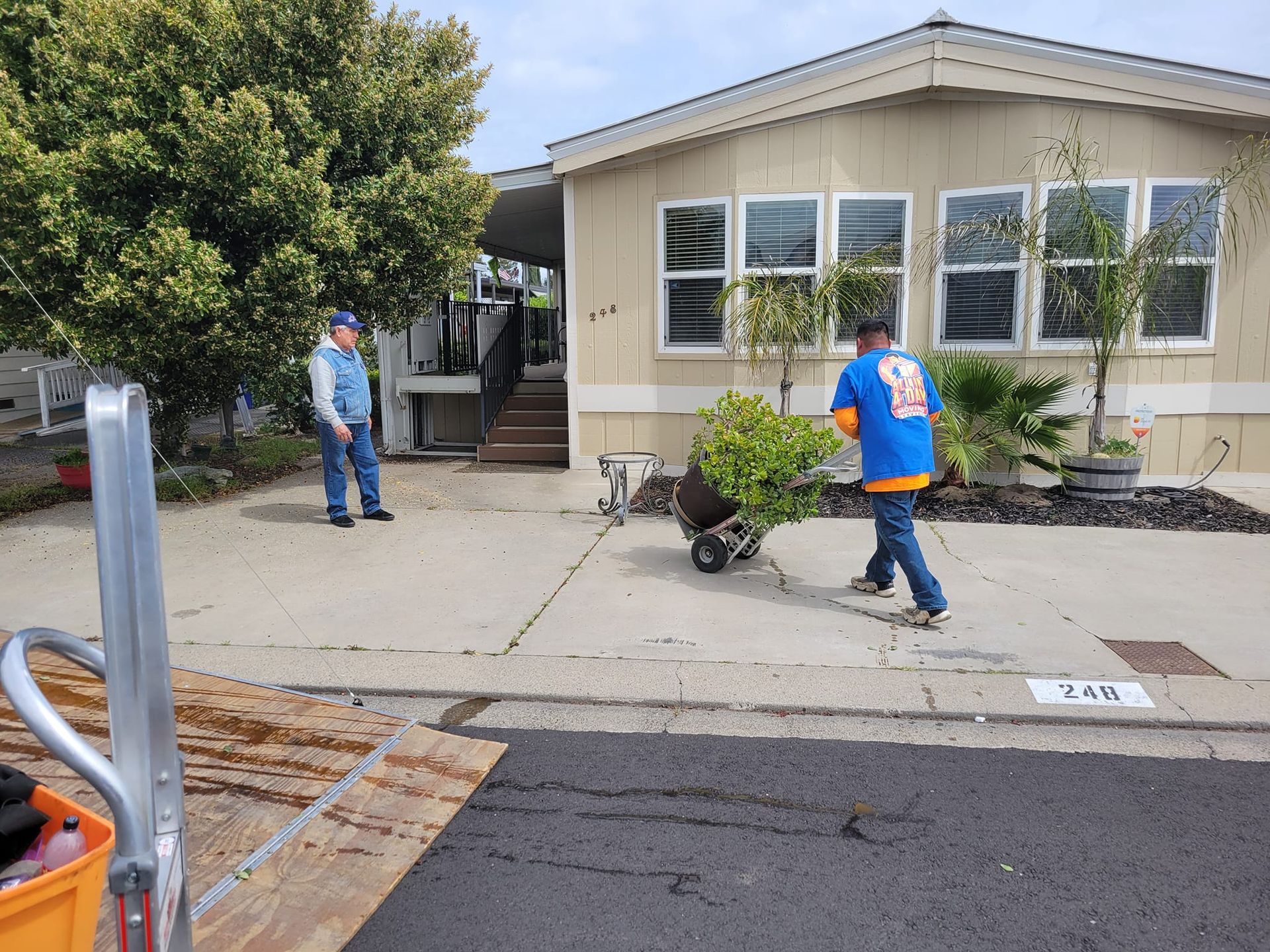 Two people move a potted shrub on a hand truck outside a mobile home on a sunny day.