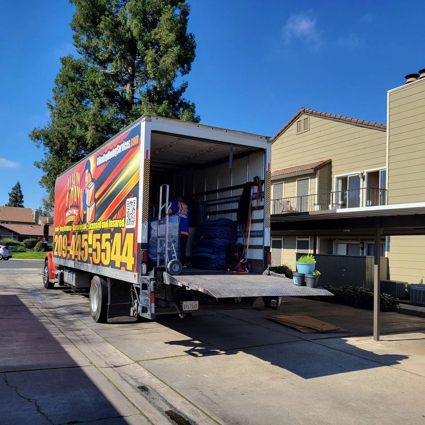 A moving truck parked at an apartment complex, with the back gate lowered and items loaded inside.