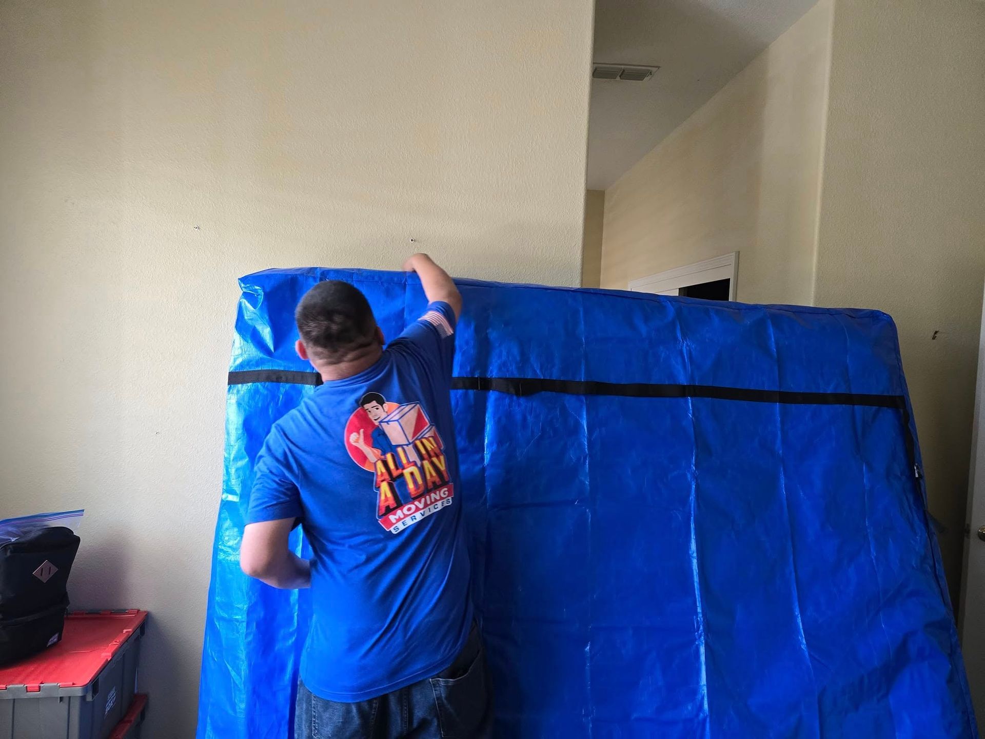 A person in a blue shirt with a company logo adjusts a large, blue protective moving cover against a beige wall.