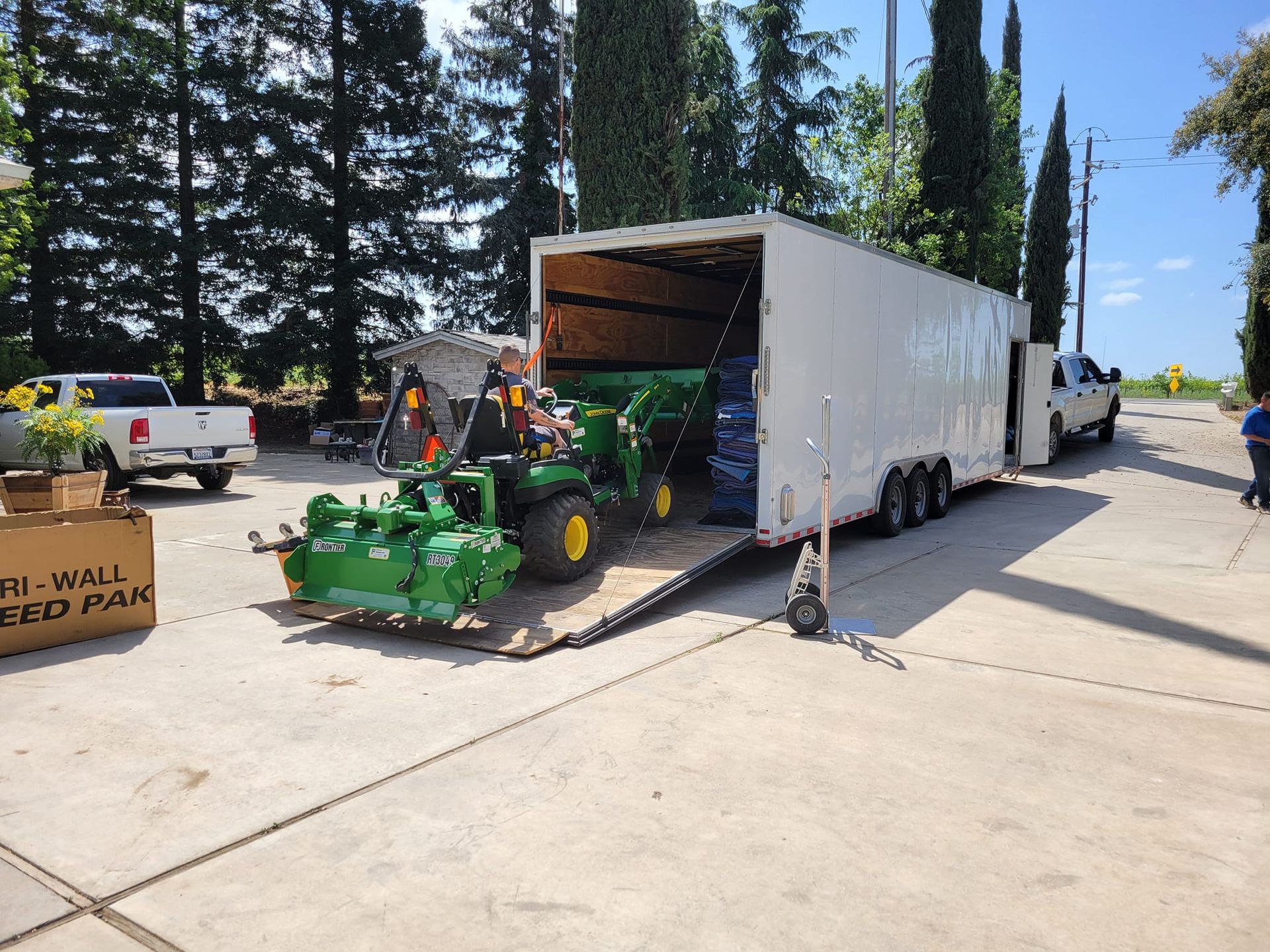 A green tractor exits a white enclosed trailer on a paved lot with parked trucks nearby.