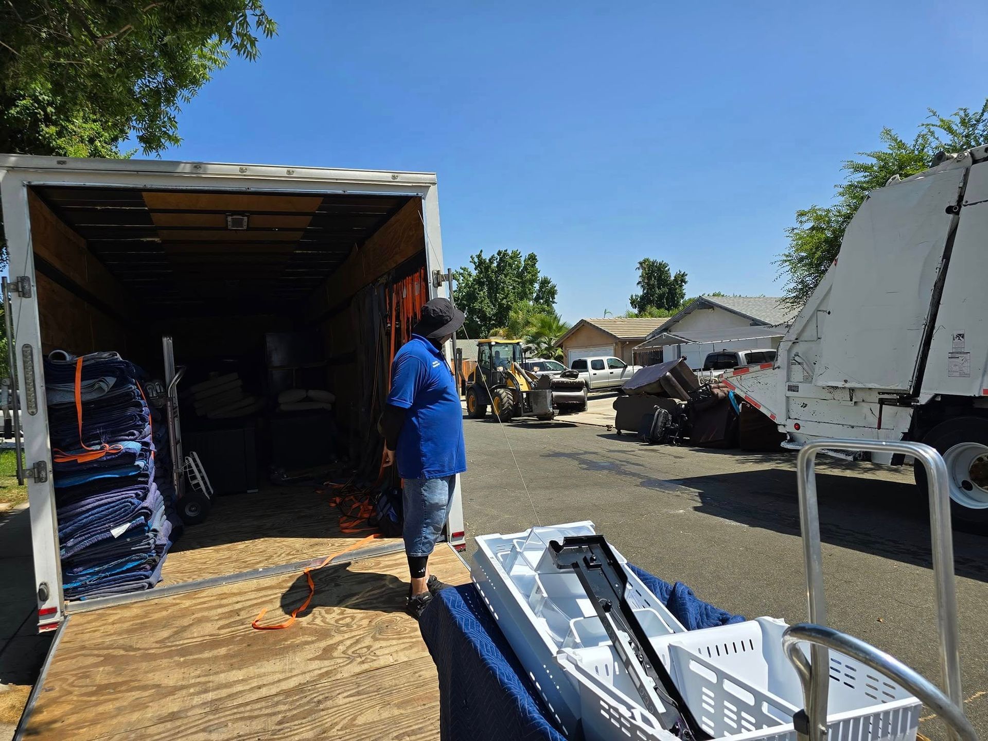 A person standing by an open moving truck at a residential site, with a heavy-duty vehicle and household items nearby.