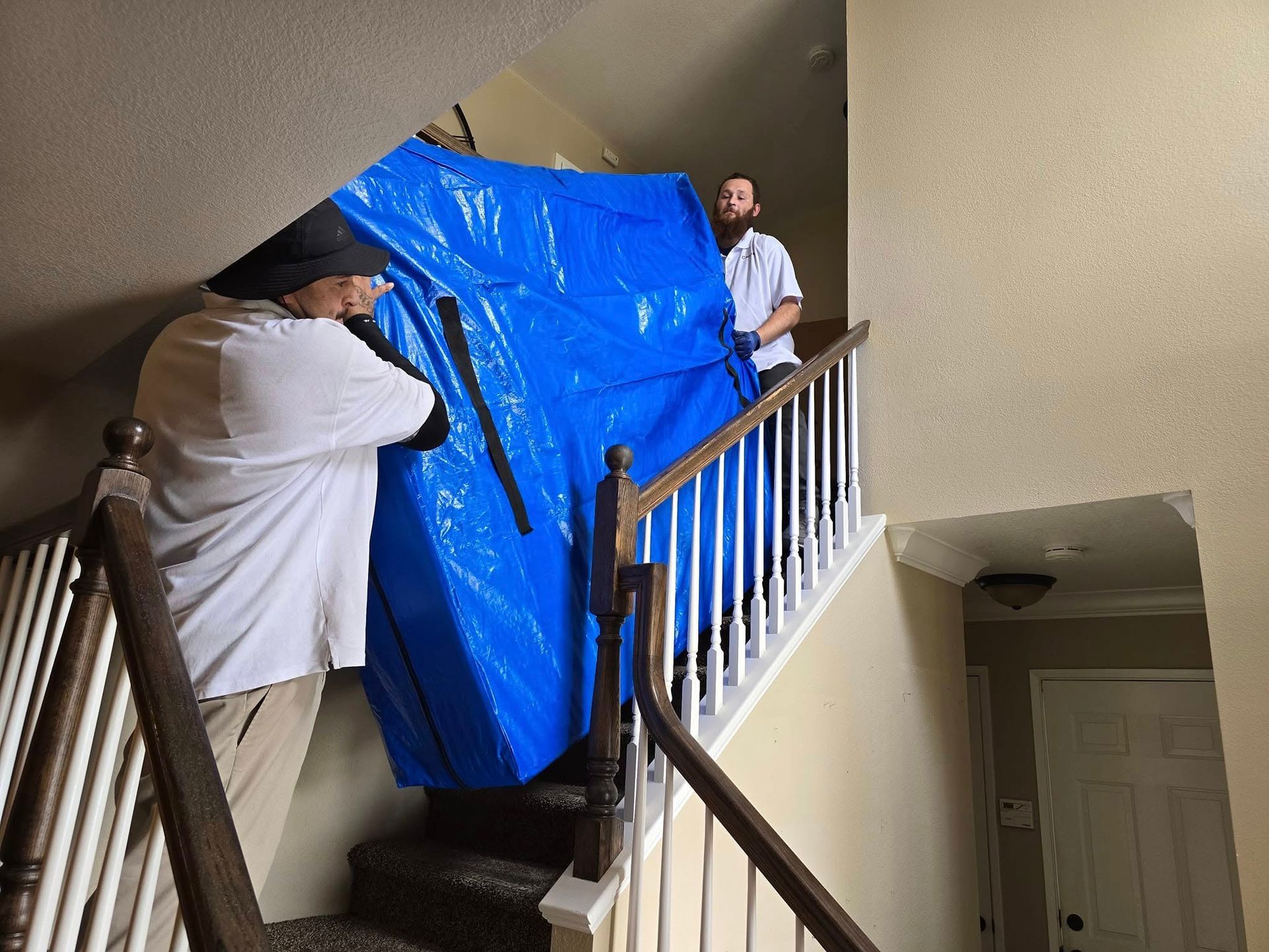 Two movers in white shirts carry a large, blue-wrapped item up a residential staircase.