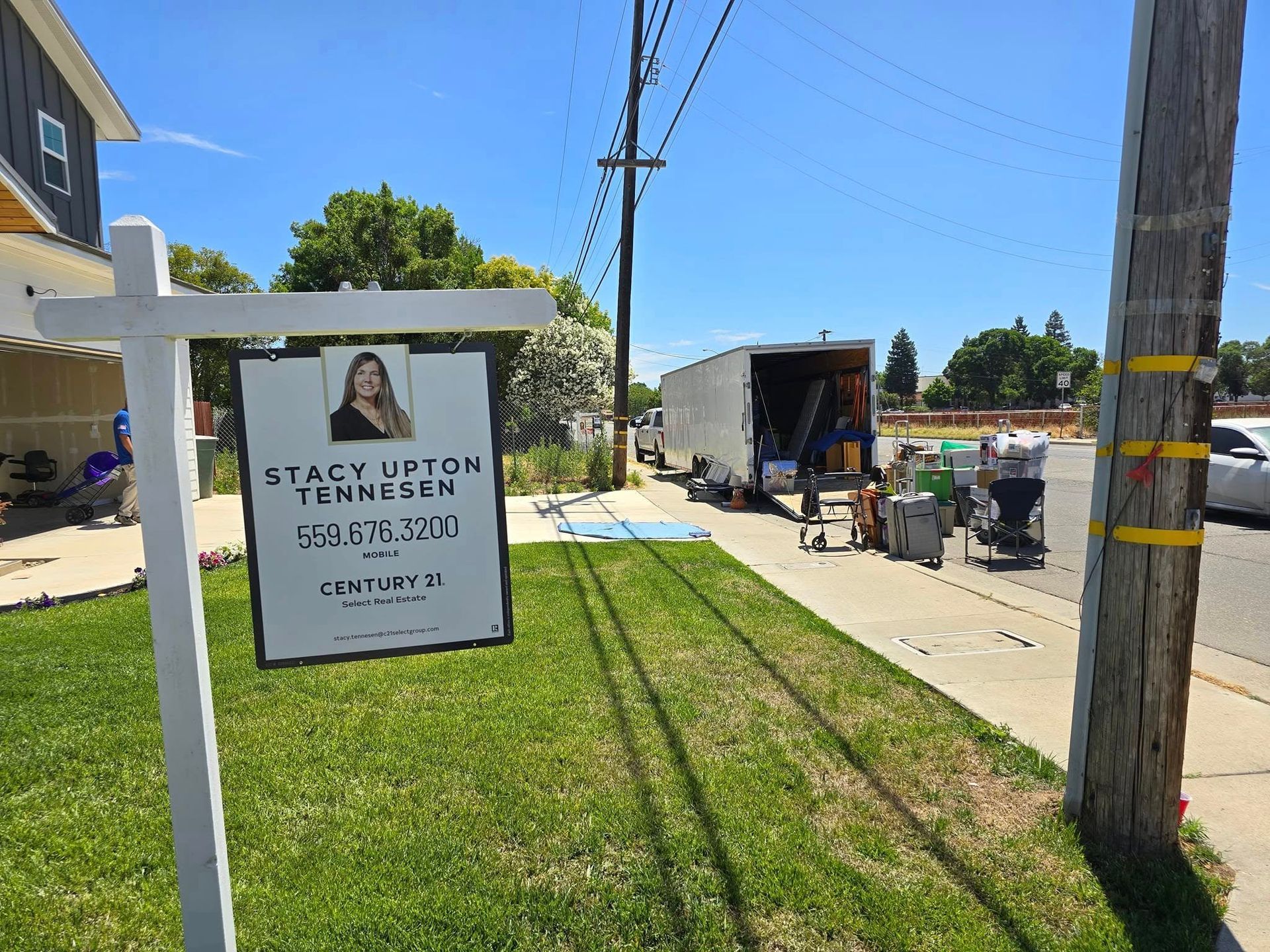 A real estate sign for Stacy Upton-Jenkson stands in a grassy yard, with a moving truck parked on the street behind it.