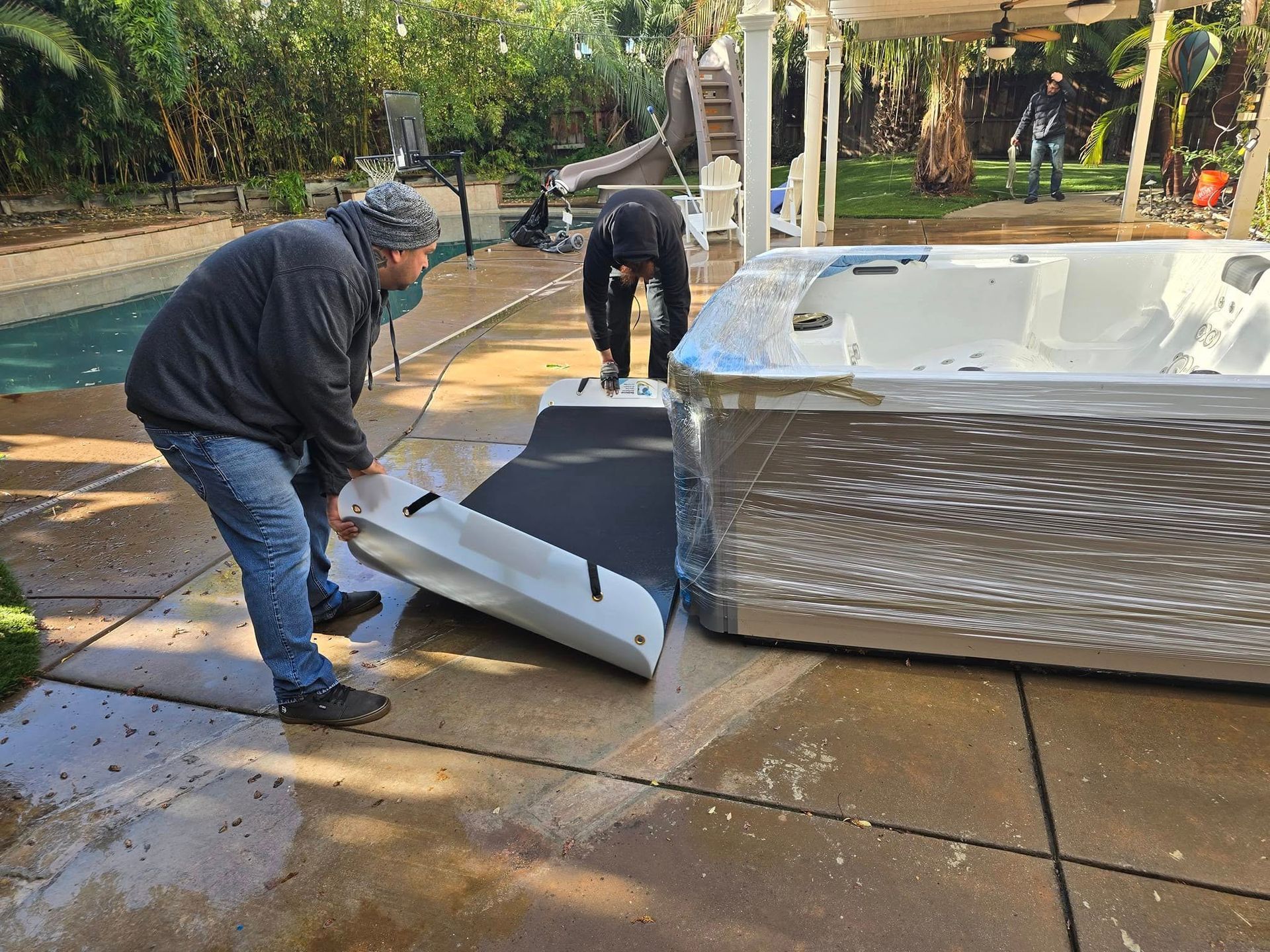 Two workers install a rectangular white cover onto a large, silver-sided hot tub on a backyard patio.